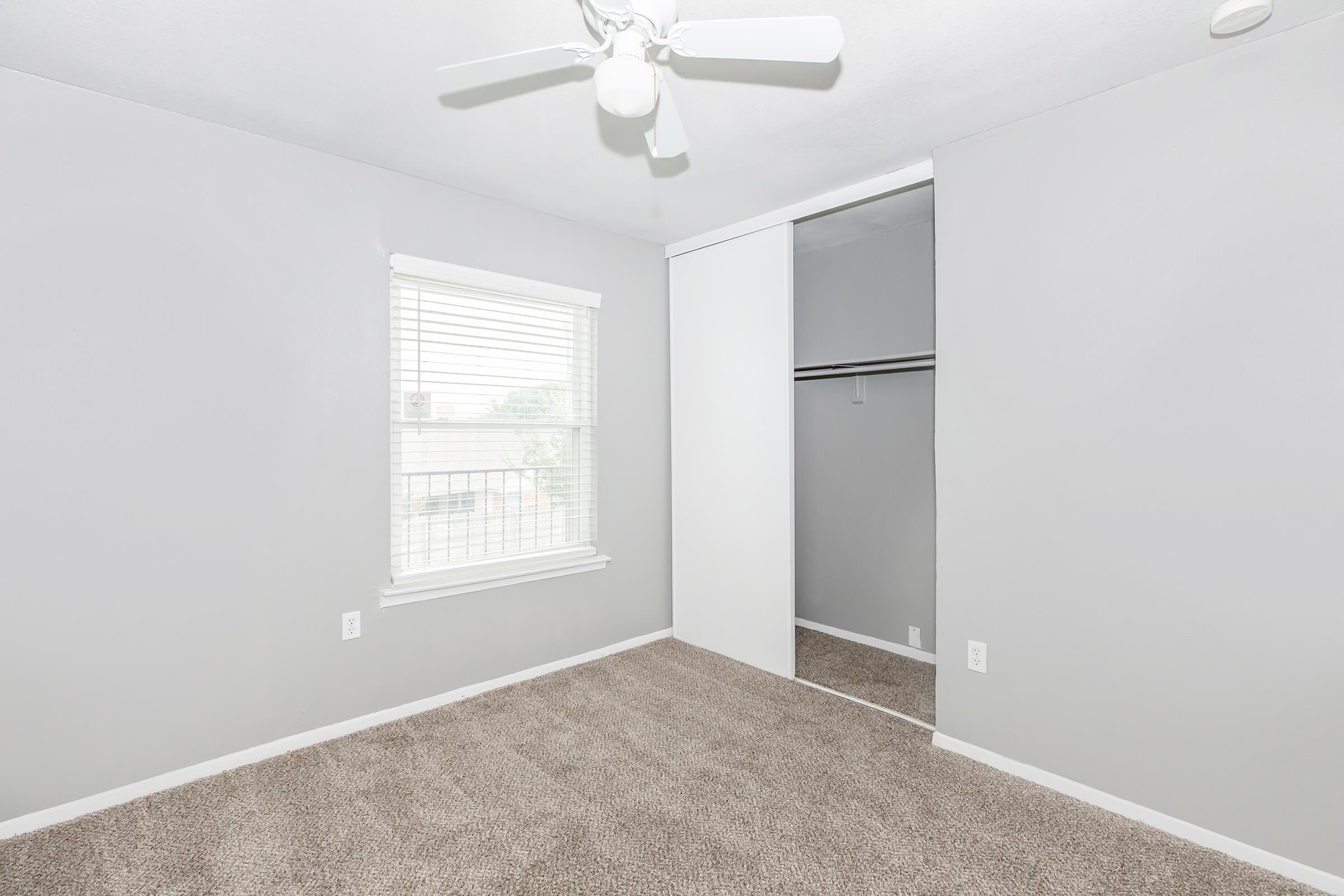 A small, empty room with light gray walls and a carpeted floor. There is a window with white blinds allowing natural light to enter. A ceiling fan is mounted above, and there is a closet with sliding doors on one side of the room. The space is clean and minimal, offering a bright and airy atmosphere.