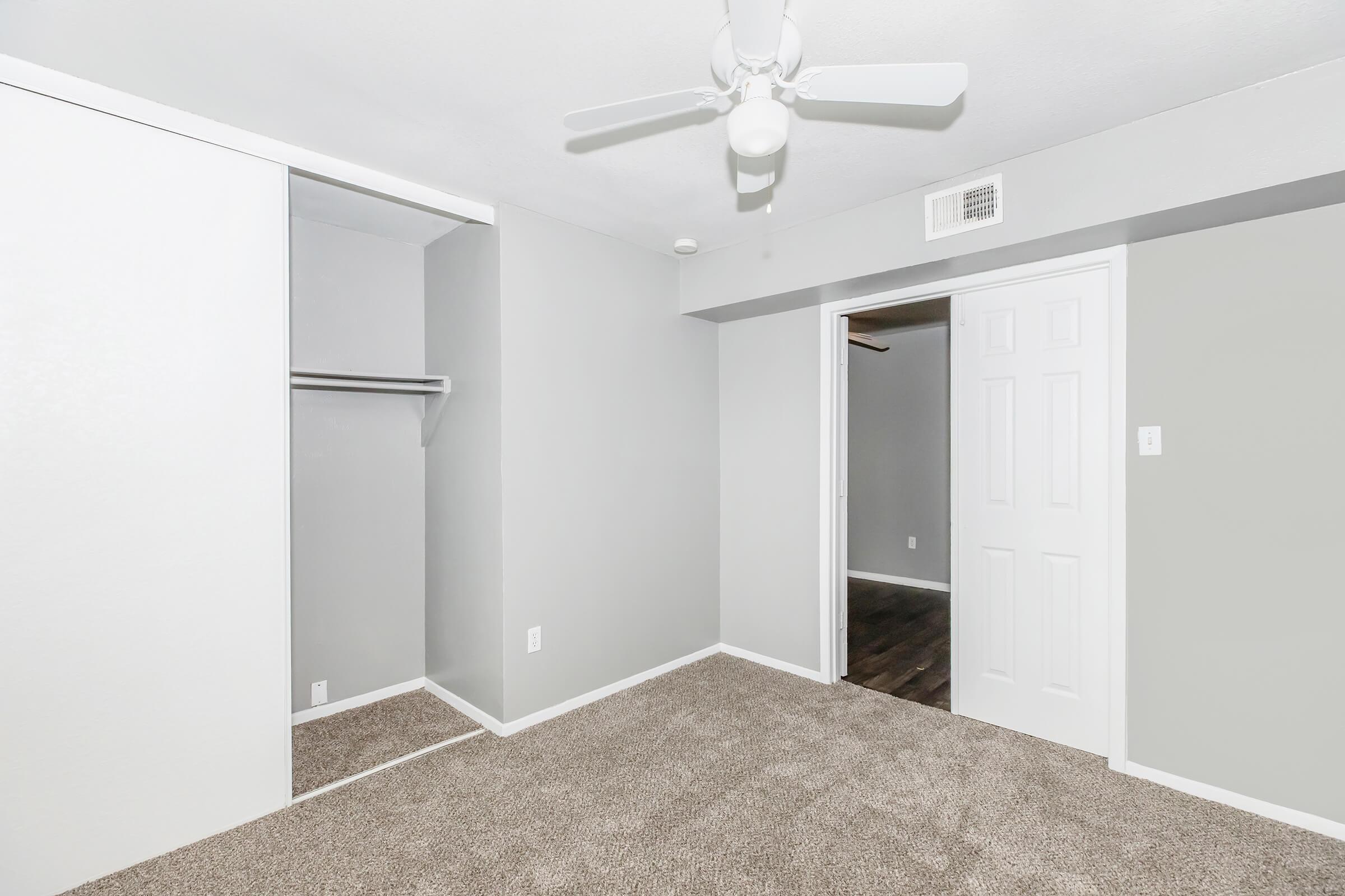 A small, empty bedroom featuring light gray walls and a ceiling fan. The room includes a closet with a sliding door on the left and a doorway leading to another room on the right. The floor is covered with carpet, creating a cozy atmosphere.
