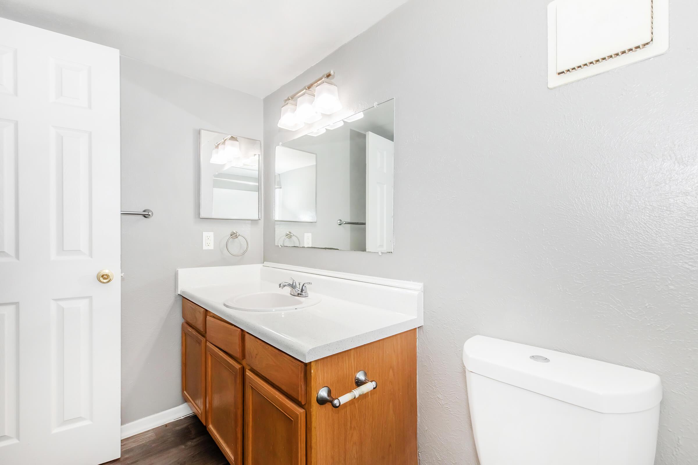 A modern bathroom featuring a wooden cabinet sink with a white countertop, a mirror above the sink, and a toilet. The walls are painted gray, and there is a light fixture with three lights above the mirror. A white door is partially visible on the left. The flooring appears to be dark wood or laminate.