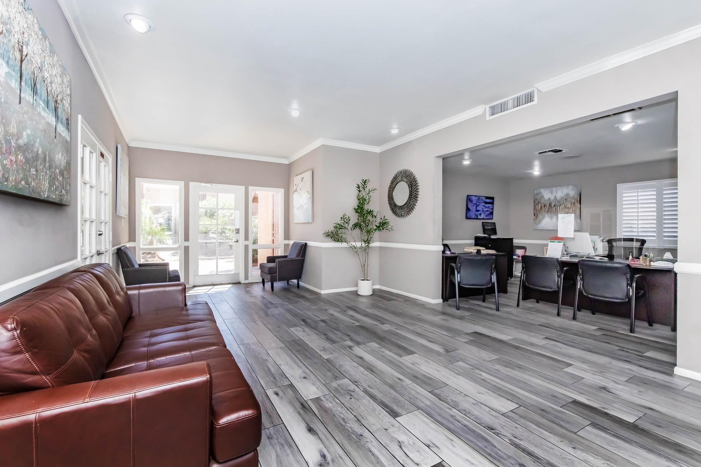 A modern reception area with a brown leather sofa, two black armchairs, and decorative plants. Light gray walls feature artwork, and large windows allow natural light. A reception desk with a computer is visible in the background, creating a welcoming atmosphere.