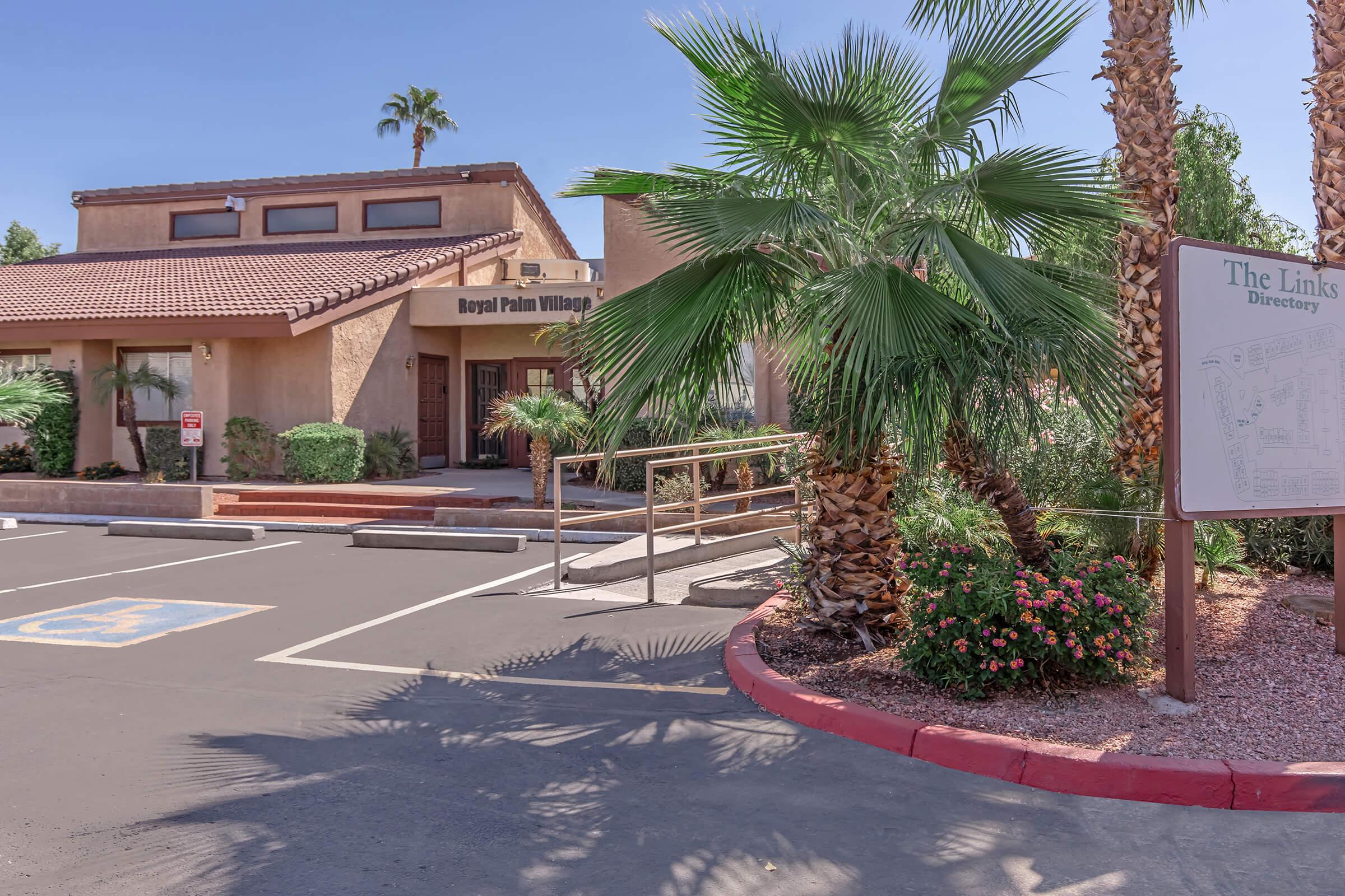 Entrance to Royal Palm Village with palm trees and a ramp leading up to the building. A directory sign for "The Links" is visible in the foreground. Accessible parking spaces are marked nearby, and the architecture features a southwestern style with a red-tiled roof.
