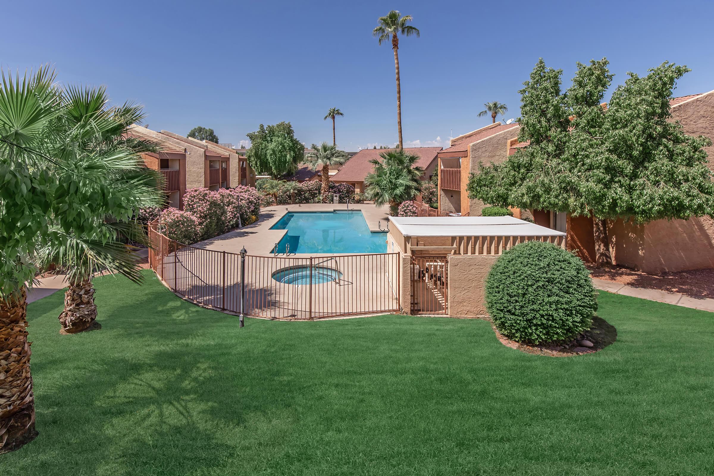 A serene view of a landscaped backyard featuring a sparkling pool surrounded by a gated area. Lush green grass, palm trees, and flowering shrubs frame the scene, with brown residential buildings in the background under a clear blue sky. A relaxing oasis perfect for outdoor enjoyment.