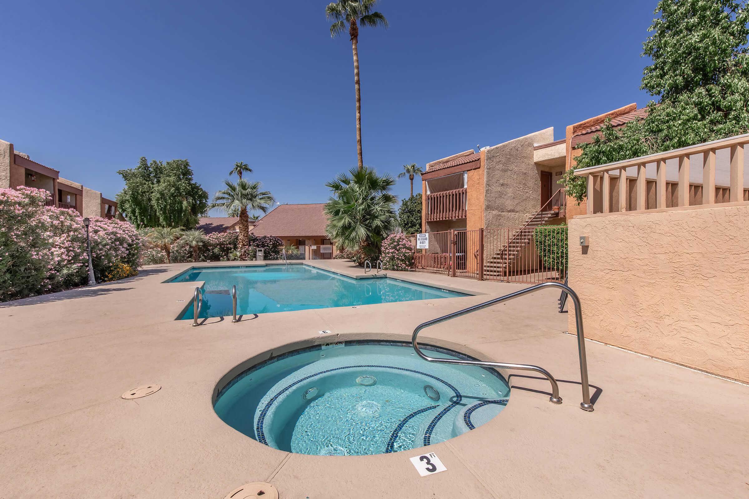A sunny outdoor area featuring a swimming pool and a hot tub. Lush greenery and palm trees surround the pools, with beige and brick-colored buildings in the background. The sky is clear and blue, creating a relaxing atmosphere. Steps leading to the upper level are visible beside the pool area.