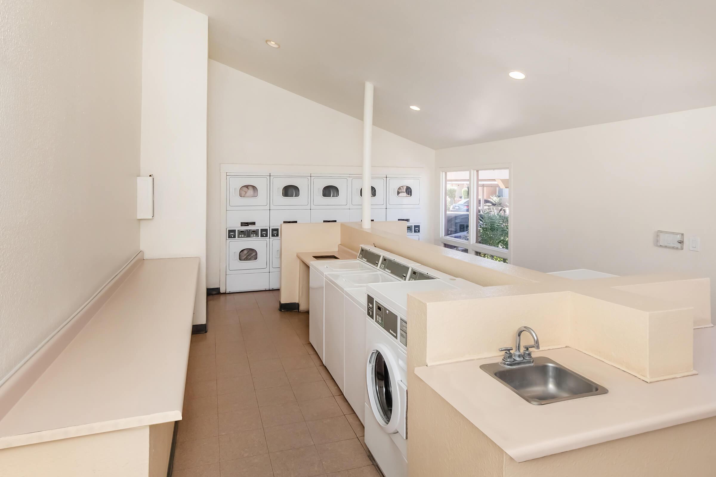 A tidy laundry room featuring white washing machines and dryers lined against the wall, a counter space with a small sink, and bright, well-lit walls. Large windows provide natural light, and the overall atmosphere is clean and organized.