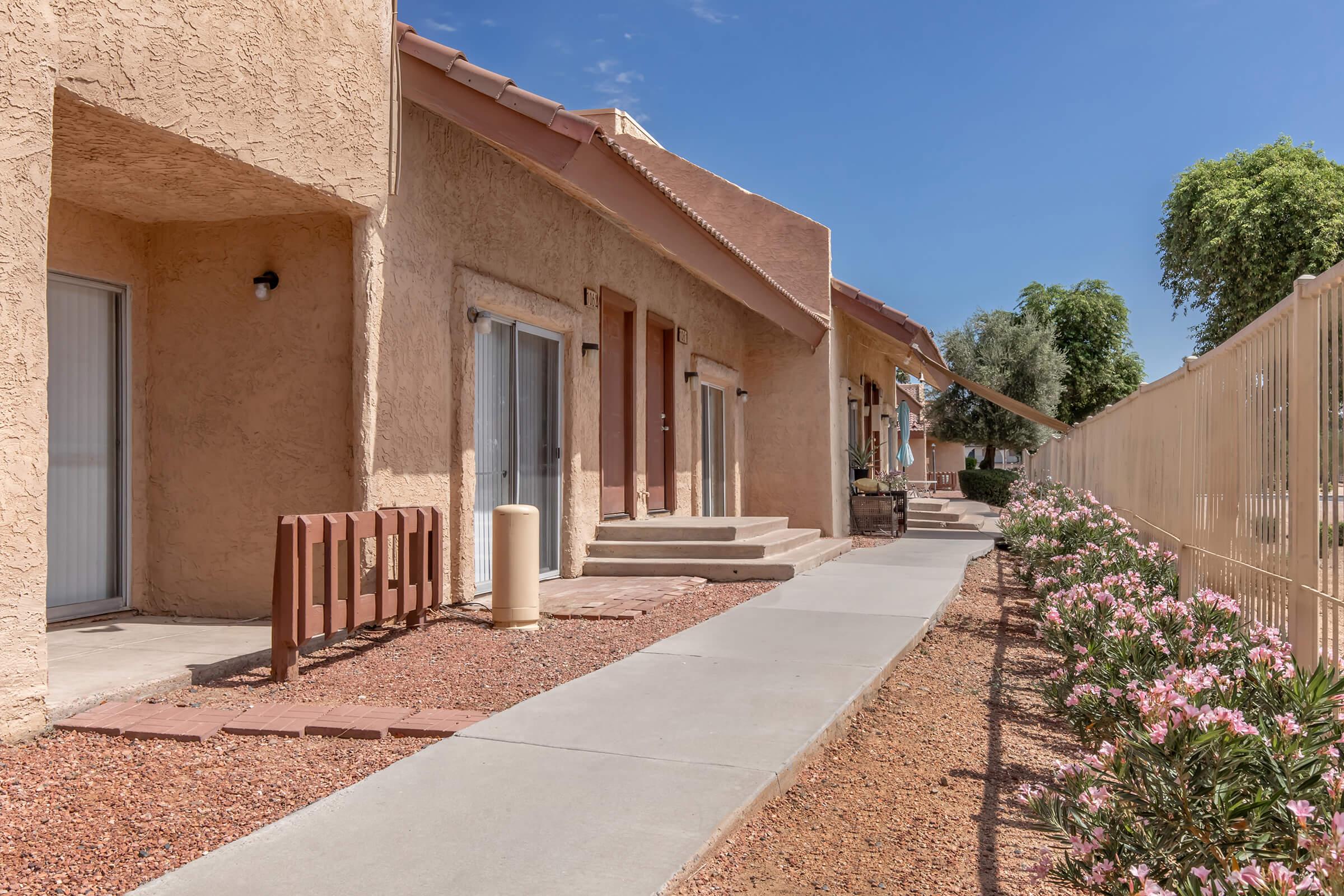 A row of modest, single-story apartment units with beige stucco exteriors. Each unit features a small porch and sliding glass doors. The pathway leading to the units is concrete, surrounded by landscaped areas with pink flowering plants and a fence on one side. The sky is blue with a few clouds.