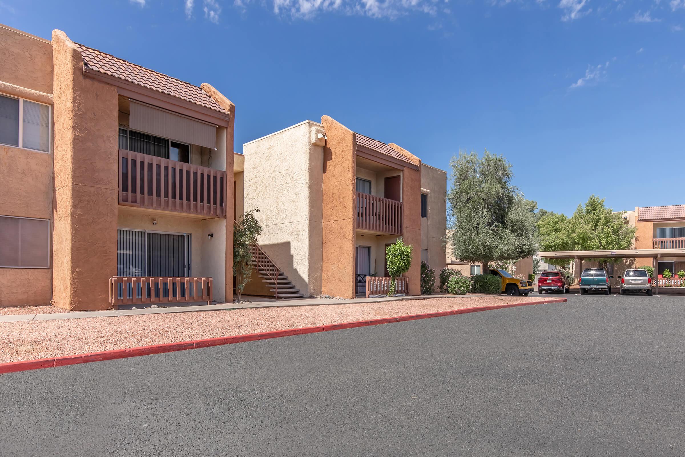 Two-story apartment buildings with a beige and brown exterior, featuring balconies and a stairway. In the foreground is a parking lot with several vehicles, bordered by landscaping and a clear blue sky. The setting is tranquil, surrounded by greenery.