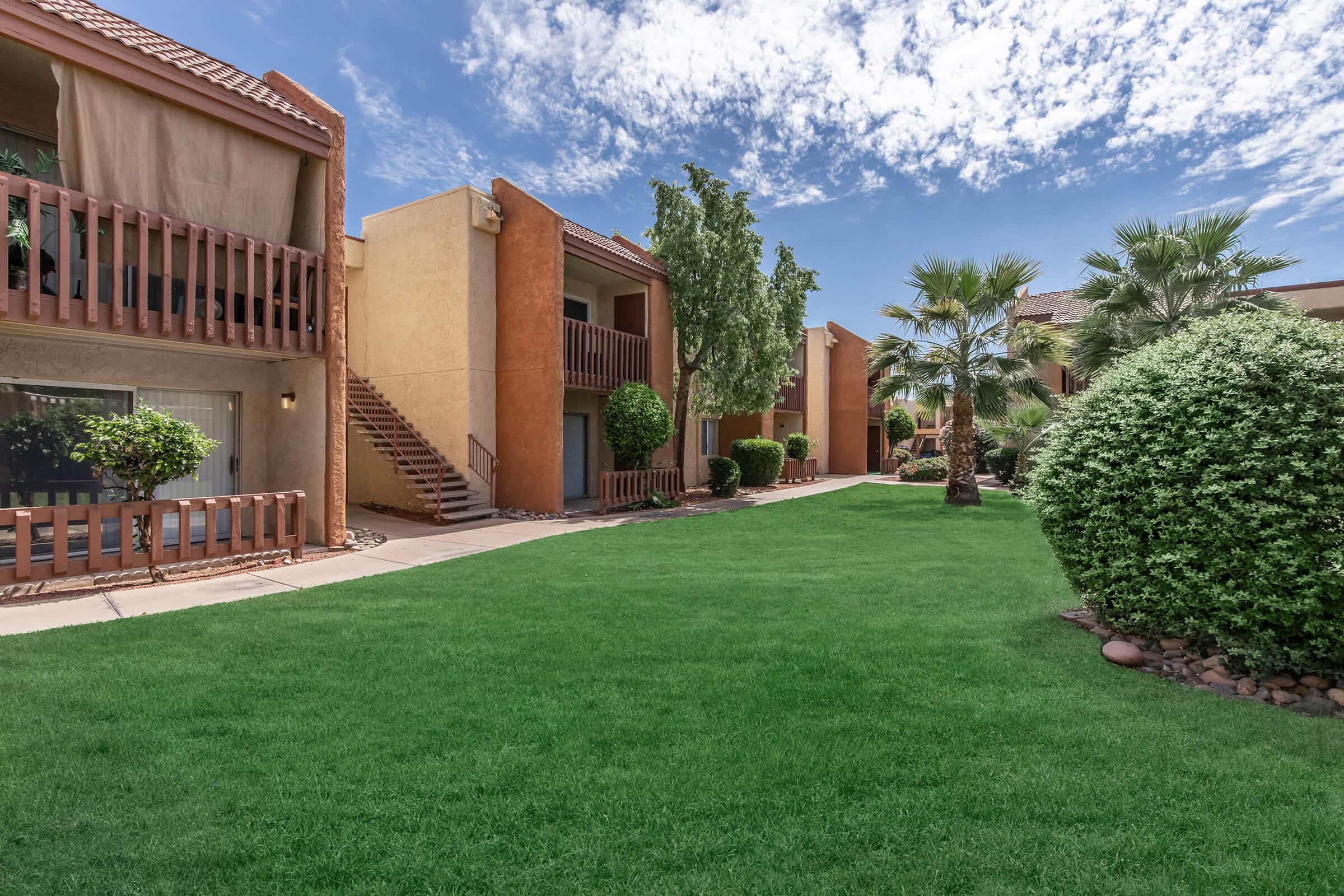 A well-maintained apartment complex featuring two-story buildings with balconies. Lush green grass and shrubs surround the pathways, and palm trees add to the tropical ambiance. The sky is clear with scattered clouds, creating a bright and inviting atmosphere.