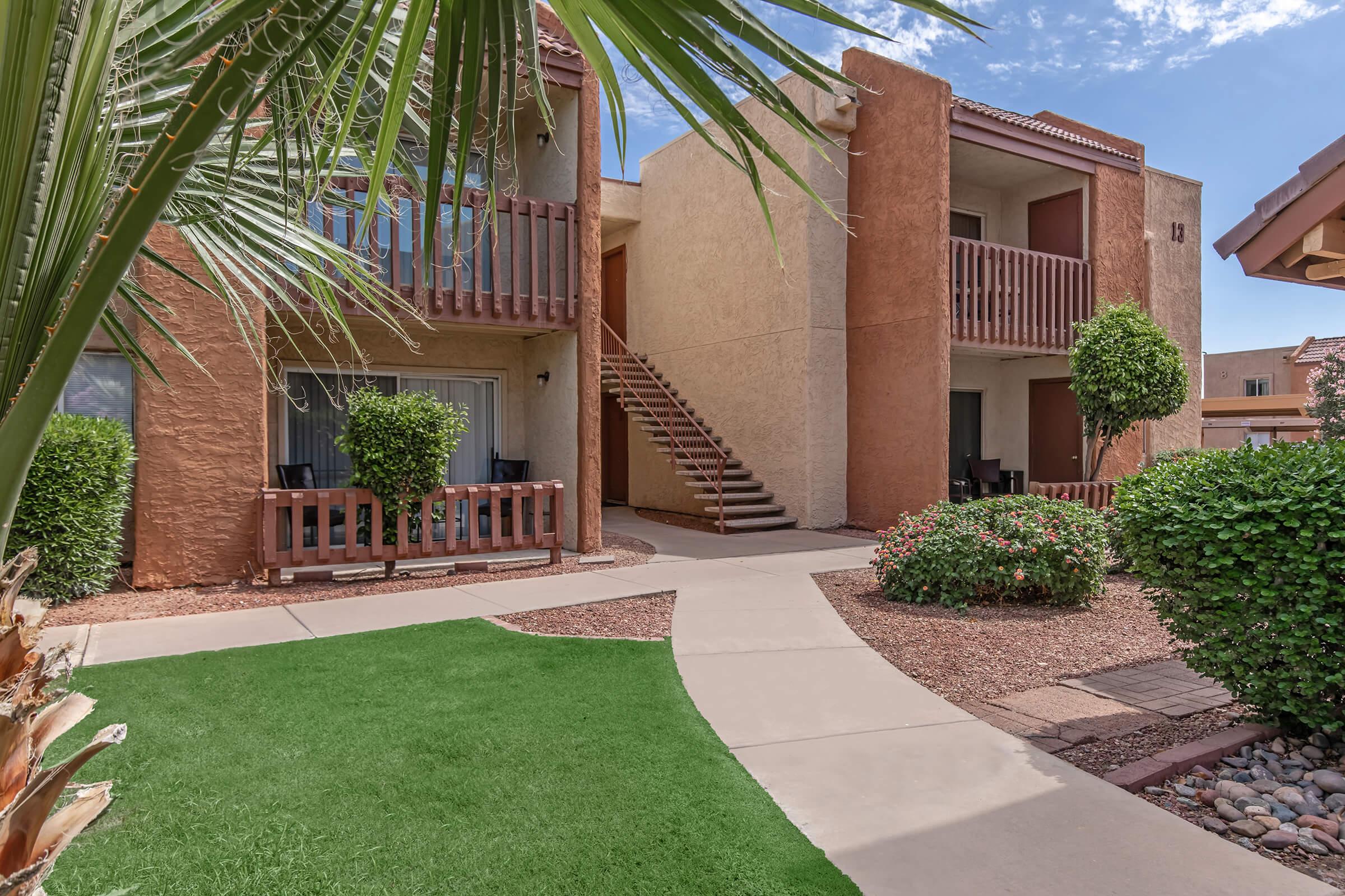 A view of a landscaped courtyard in an apartment complex featuring two-story buildings with balconies, a winding path, and greenery, including plants and shrubs, under a clear blue sky.