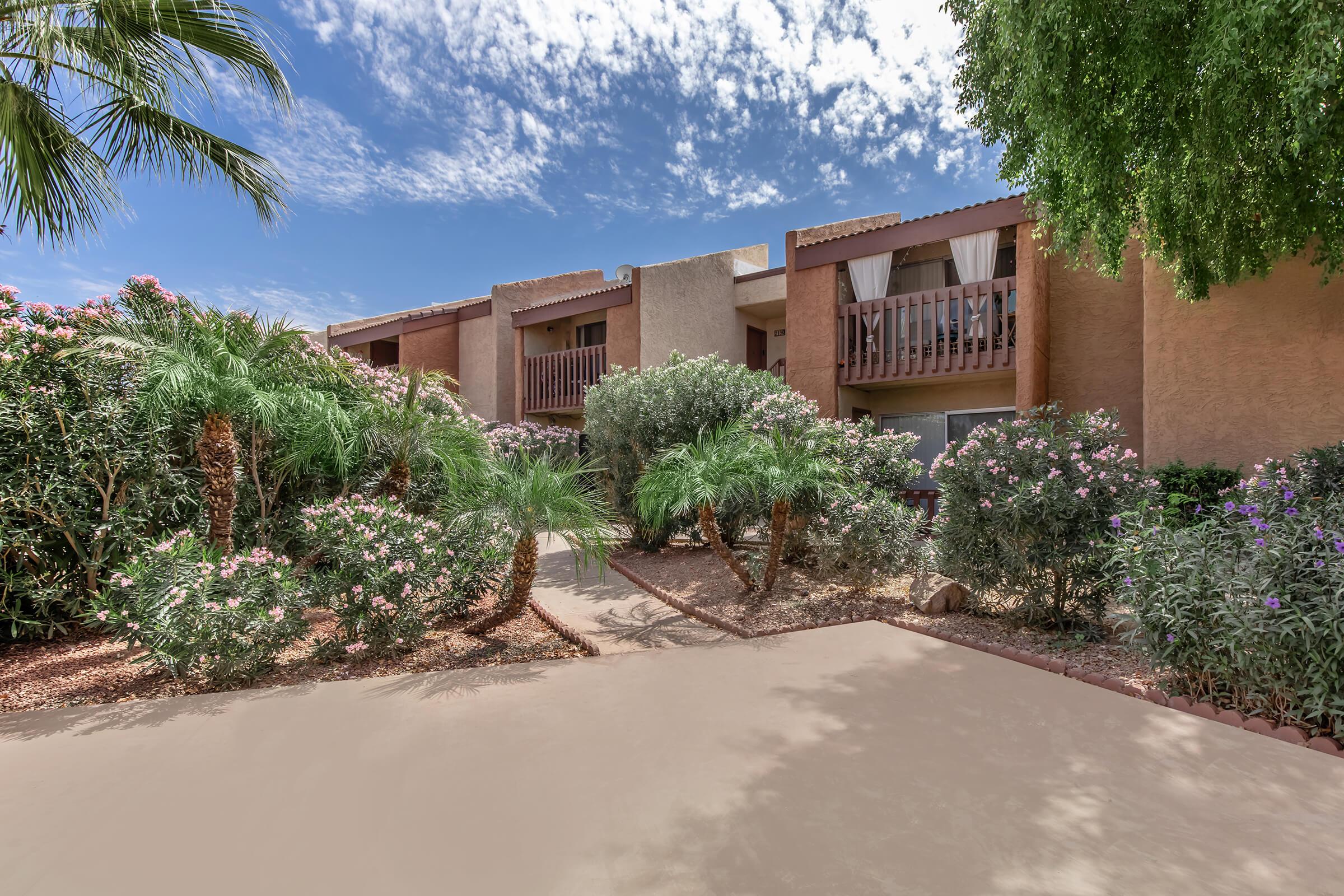 A landscaped courtyard featuring lush palm trees and flowering bushes, leading to a multi-level building with balconies. The sky is bright with scattered clouds, creating a warm and inviting atmosphere. The pathway is clear, providing access to the building's entrance.