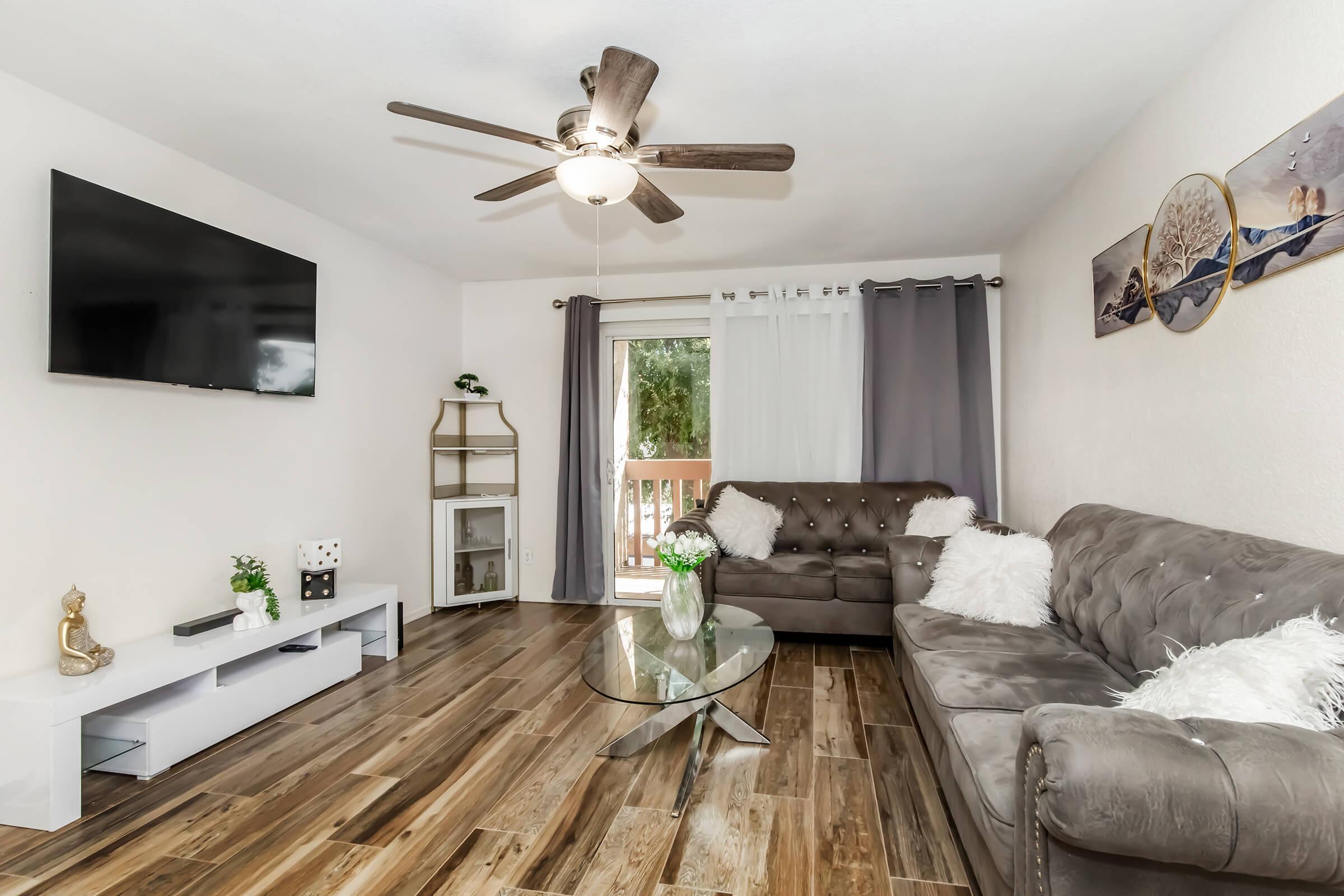 A cozy living room featuring a brown tufted sofa and a modern glass coffee table. A wall-mounted TV is on the left, and a small shelf holds decorative items. Large windows with curtains allow natural light, and a sliding door opens to a balcony. Decorative touches include a vase of flowers and a small Buddha statue.