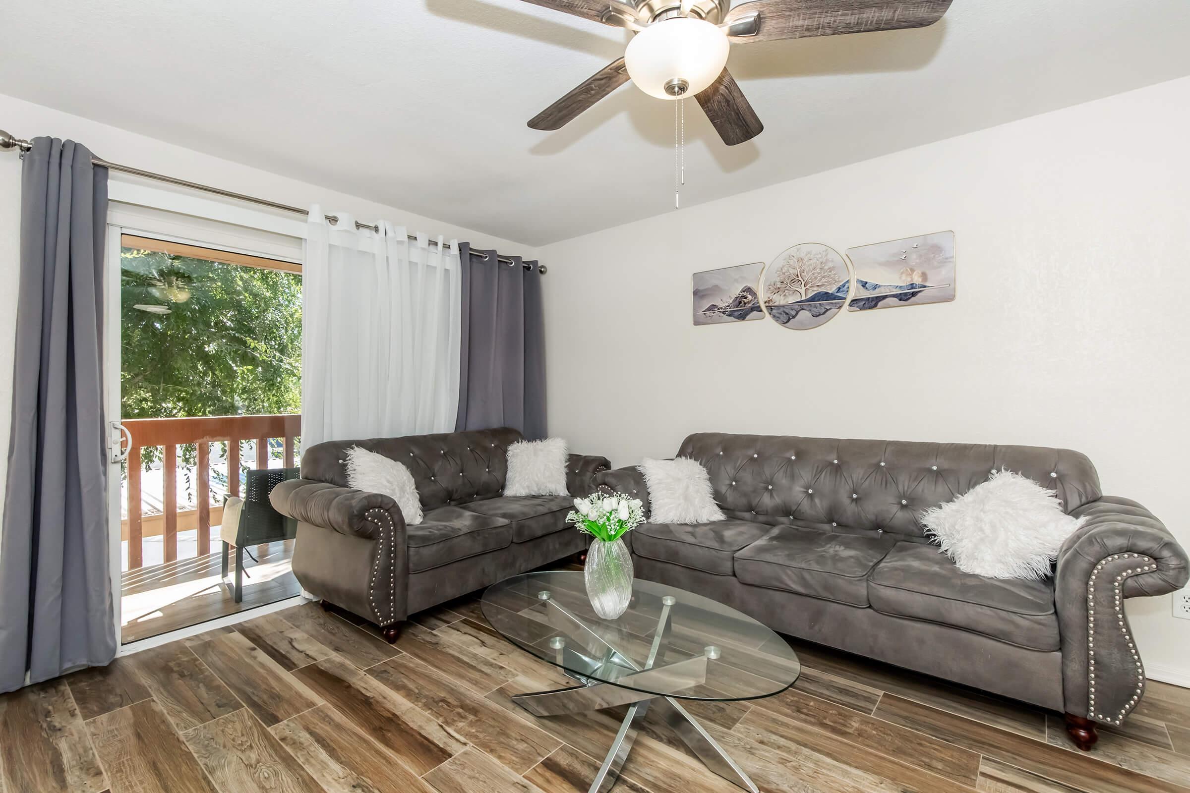 A cozy living room with two gray tufted sofas adorned with white pillows. A glass coffee table sits in the center on wooden flooring. Light curtains complement the dark ones, and a ceiling fan hangs above. A sliding door leads to a balcony with greenery visible outside, enhancing the space's inviting atmosphere.