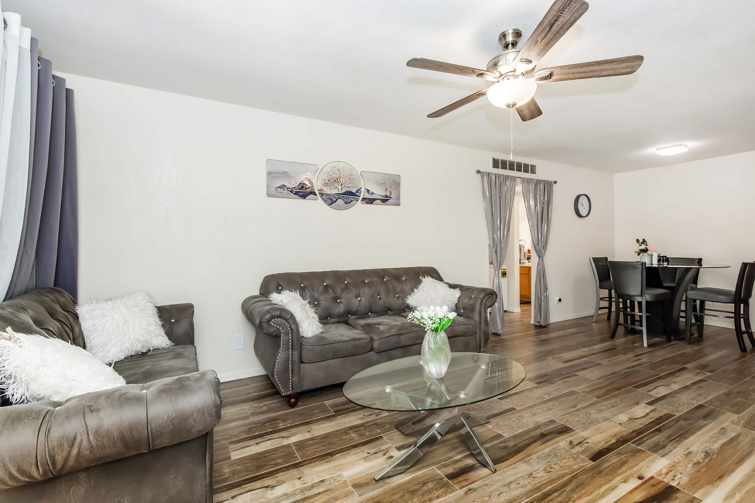 A cozy living room featuring two gray sofas with white throw pillows, a glass coffee table, and a ceiling fan. The walls are painted light colors, adorned with wall art. There are sheer curtains and a dining area with dark chairs in the background, enhancing the inviting atmosphere.