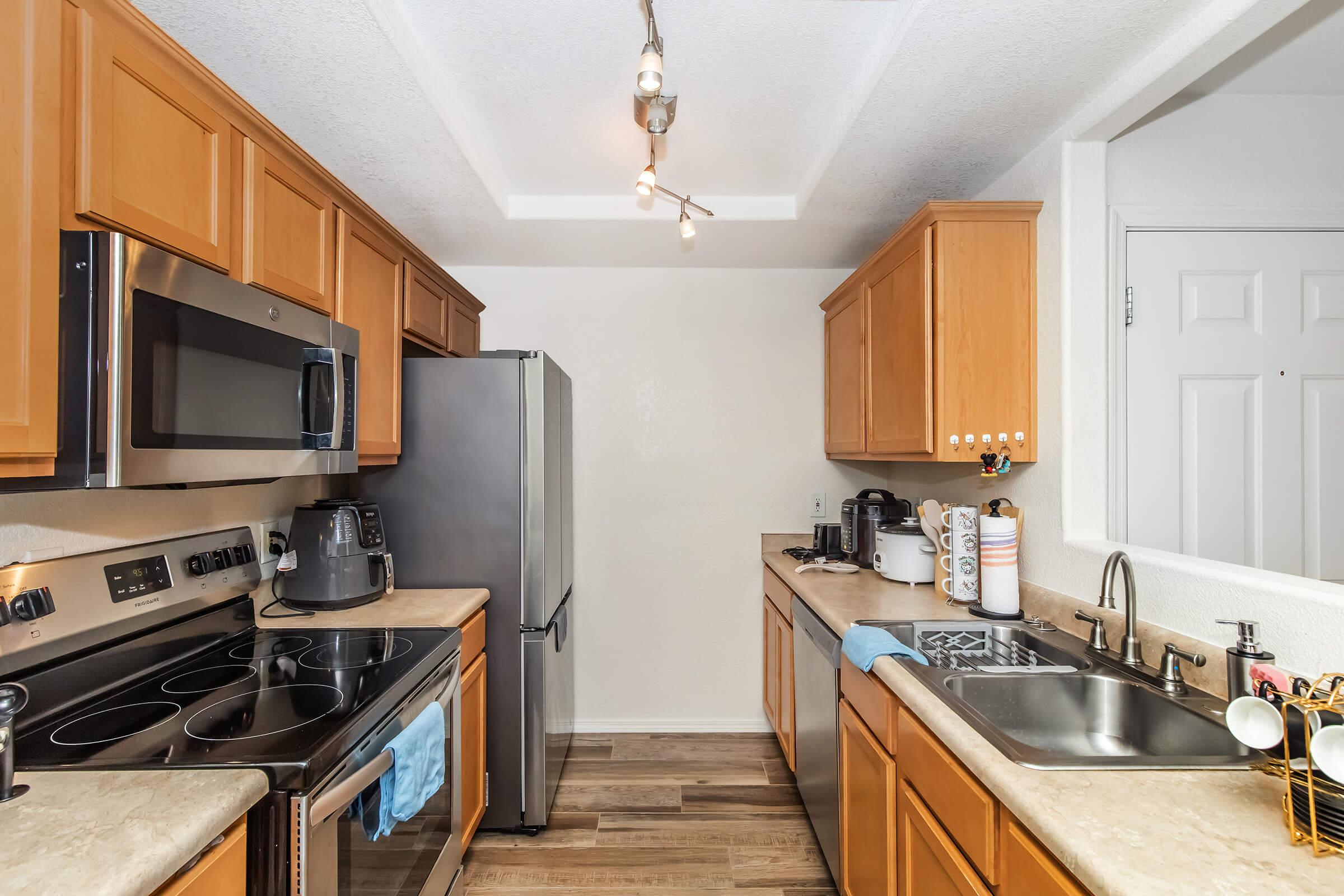 Modern kitchen with light wood cabinets, stainless steel appliances including a refrigerator and oven, and a dual sink. The countertop is beige, featuring a blue dish towel and kitchen gadgets. Soft lighting is provided by track lights along the ceiling, and the walls are painted white.