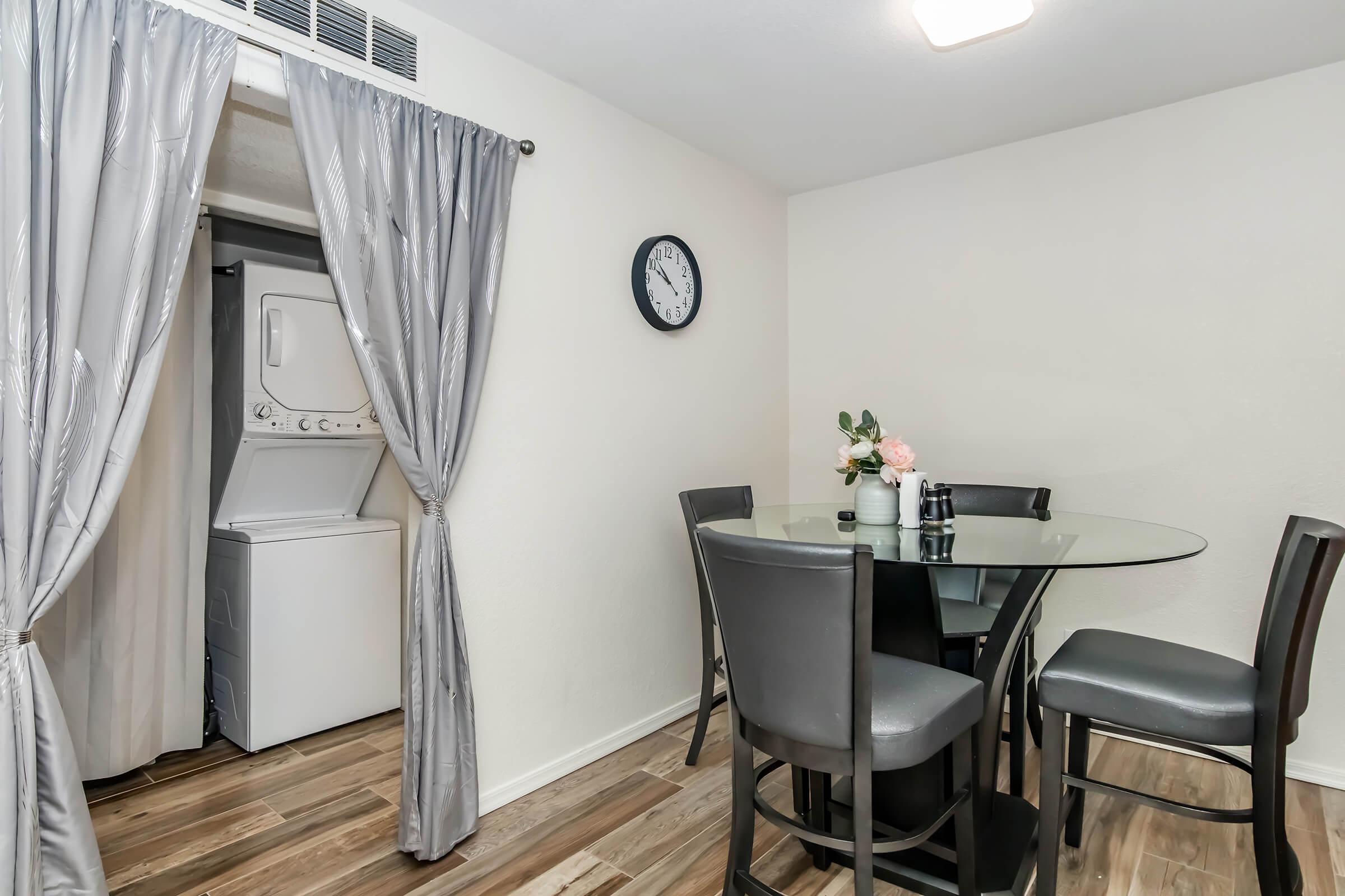 A modern dining area featuring a round glass table with four black chairs, a vase of flowers, and a wall clock. In the background, a set of curtains partially reveals a laundry area with a washer and dryer. The flooring is wood-like, and the walls are light-colored, creating a clean, contemporary look.