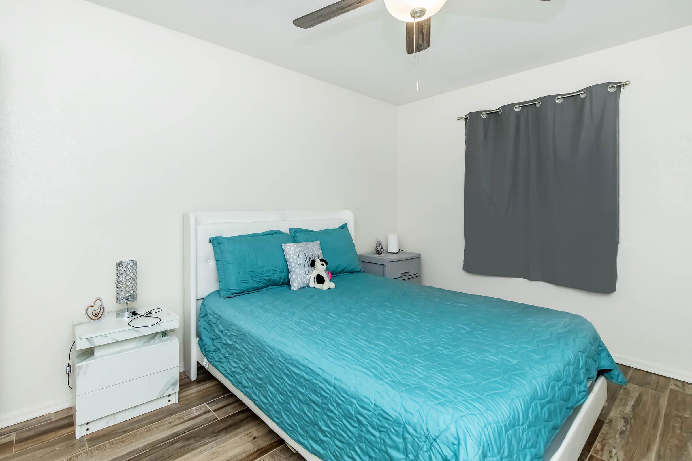 A neatly arranged bedroom featuring a white bed with a light blue quilt and matching pillows. A small teddy bear sits on the bed. There's a gray curtain hanging by a window, a nightstand with a lamp and a phone charger beside the bed, and wood-look flooring.