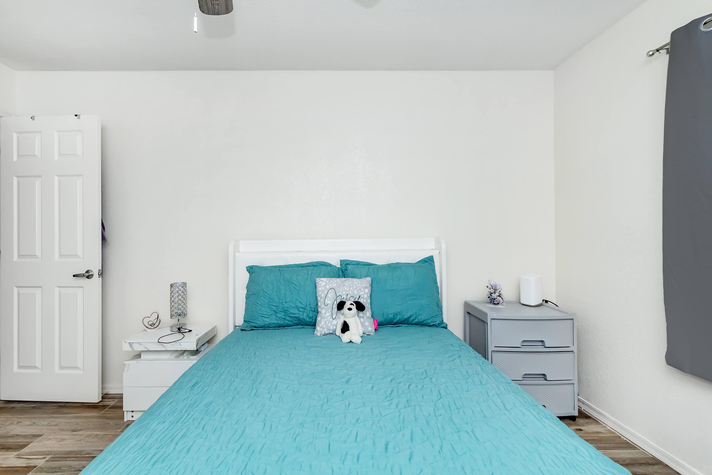A cozy bedroom featuring a light blue bedspread with two pillows, a stuffed dog on the bed, a nightstand with decorative items, and a gray dresser. The room has a light-colored wall and a door leading to another space, with a fan on the ceiling and a window covered by light gray curtains.