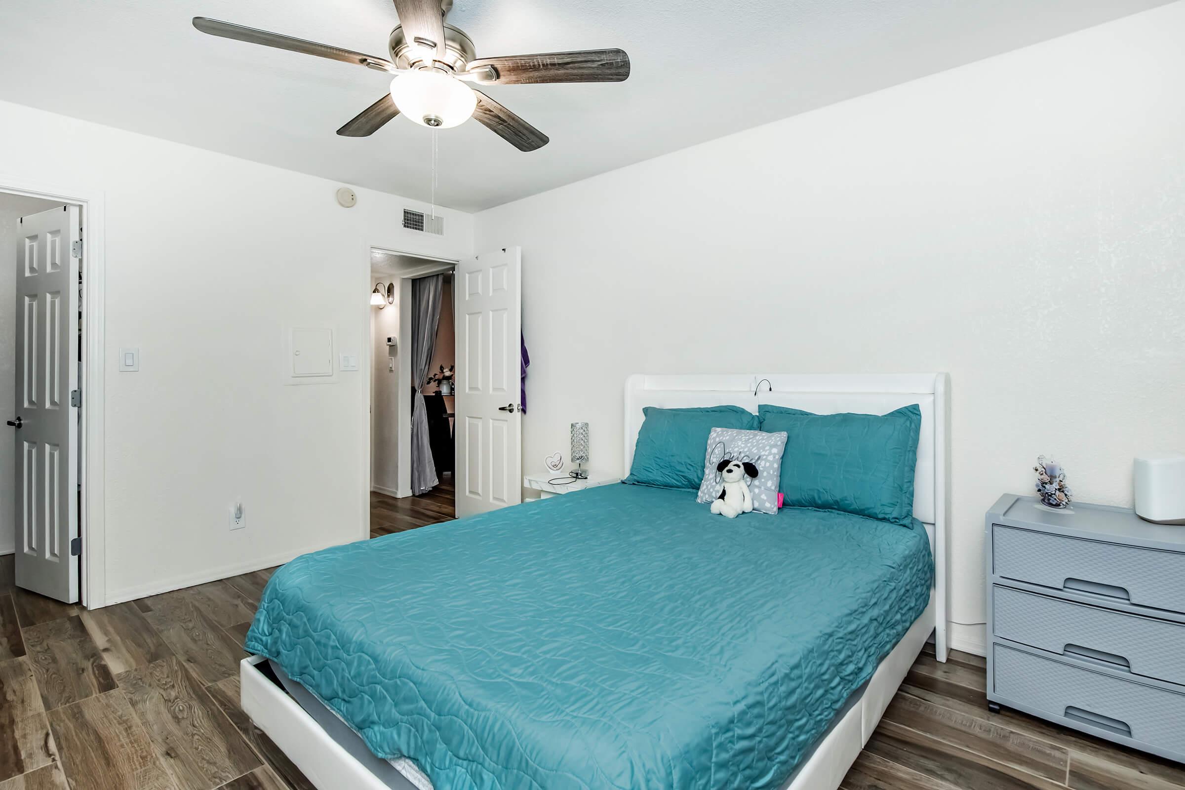 A bright and tidy bedroom featuring a queen-sized bed with a turquoise comforter and a plush toy on top. The room has a light wood floor, a ceiling fan, and neutral-colored walls. A closed closet and a light grey nightstand are visible, along with an open door leading to another space.
