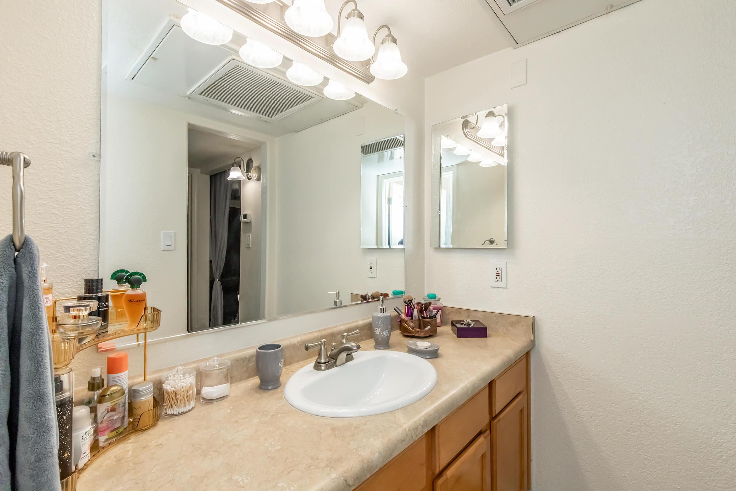 A clean bathroom with a light-colored countertop and a white sink. The space features two mirrors, a set of five light fixtures above, and various toiletries arranged neatly. Towels hang on a rack, and small containers with personal items are placed on the countertop. The walls are painted in a light shade.