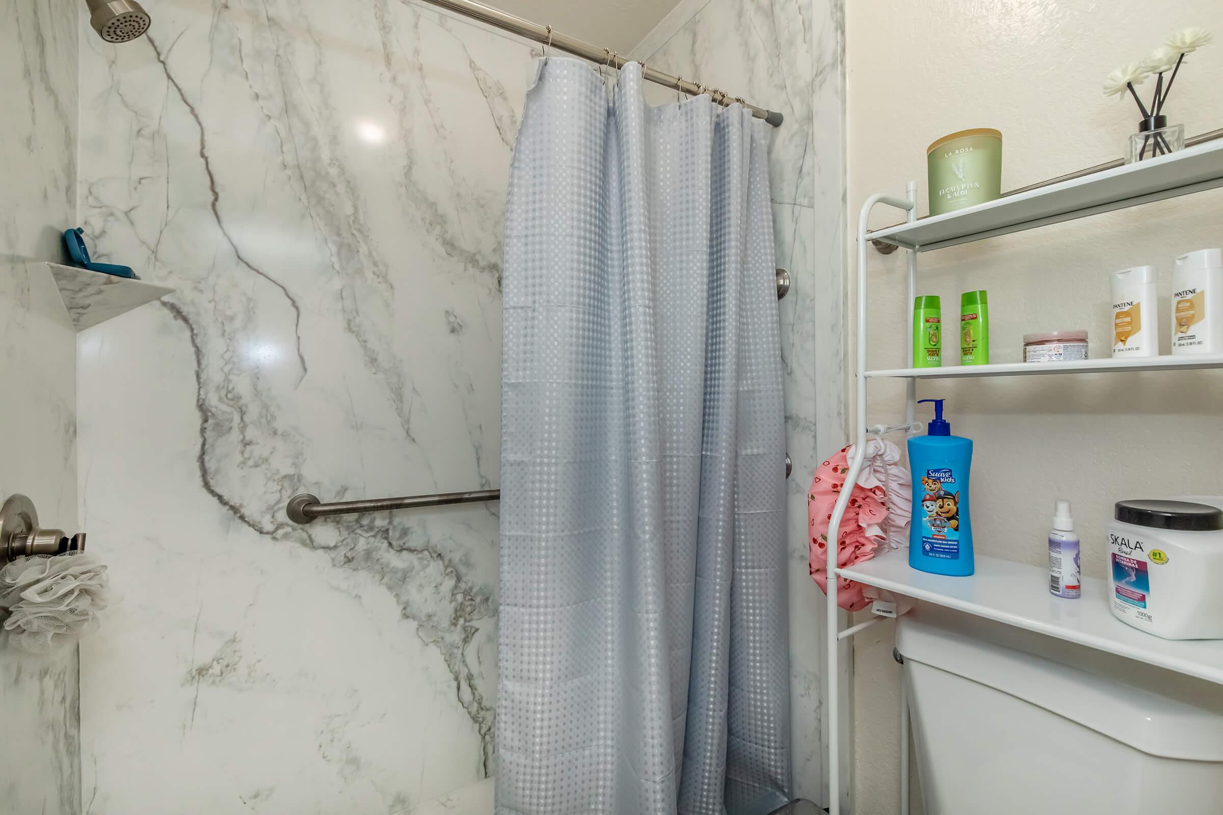 A modern bathroom shower area featuring a gray patterned shower curtain, marble walls, and a small shelf with various toiletries including shampoo and body wash. A toilet can be seen in the corner, and the overall design is clean and contemporary.