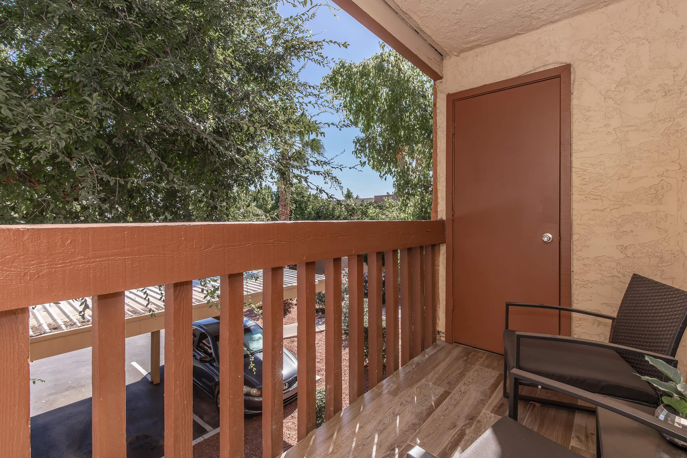 A balcony with a wooden railing, featuring a chair and a small table. The view shows a nearby parking area and trees, with a door leading into an interior space. Bright daylight illuminates the scene, creating a warm and inviting atmosphere.