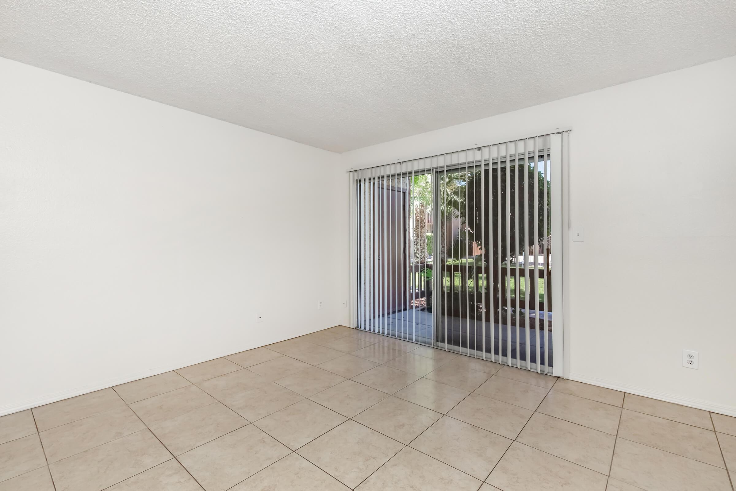 A spacious room with white walls and tiled flooring. A set of vertical blinds covers a large sliding glass door that opens to a patio or backyard area, allowing natural light to enter the space. The overall ambiance is bright and inviting.