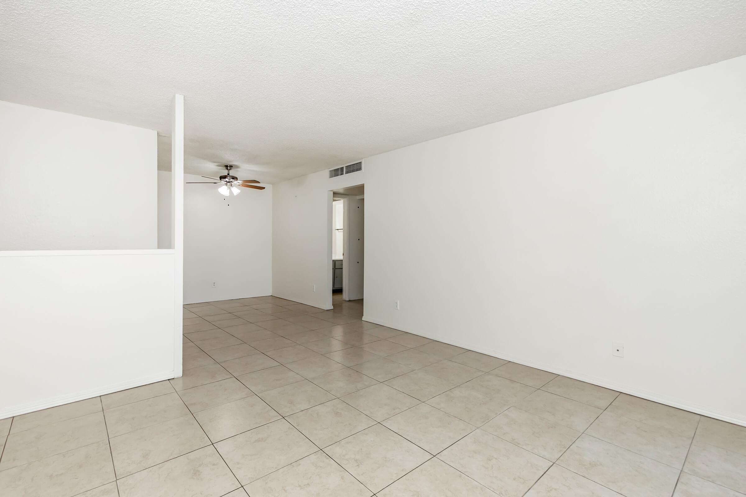 A spacious, empty living area with white walls and light-colored tile flooring. A ceiling fan is visible in the corner, and a partial wall separates the living space from a hallway leading to another room. Natural light illuminates the area, creating a bright and airy atmosphere.