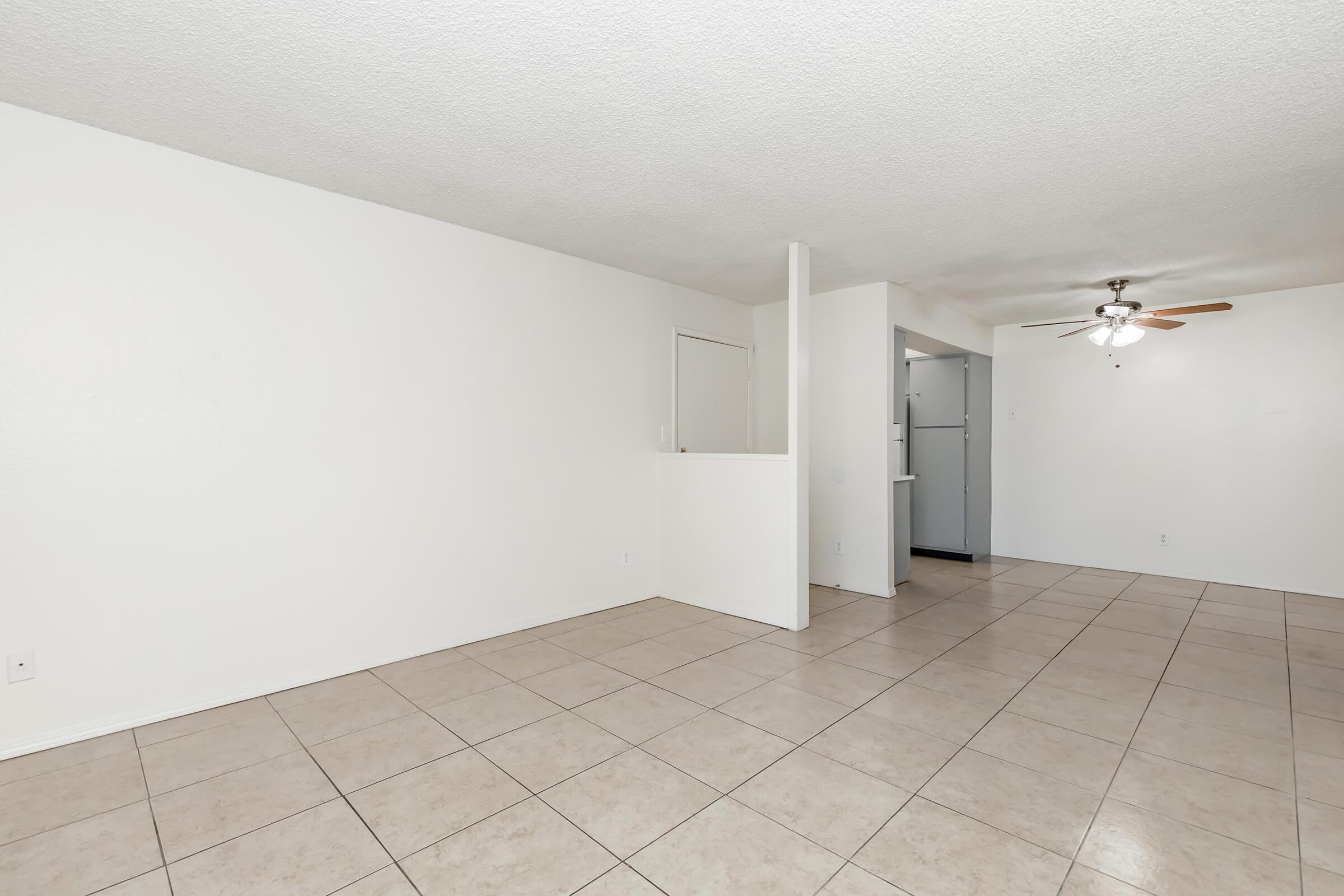 Empty living space with light-colored walls and tiled flooring. A ceiling fan is visible, and there's a partial wall separation leading to a kitchen area. The room is bright and spacious, featuring a clean and minimalist design.