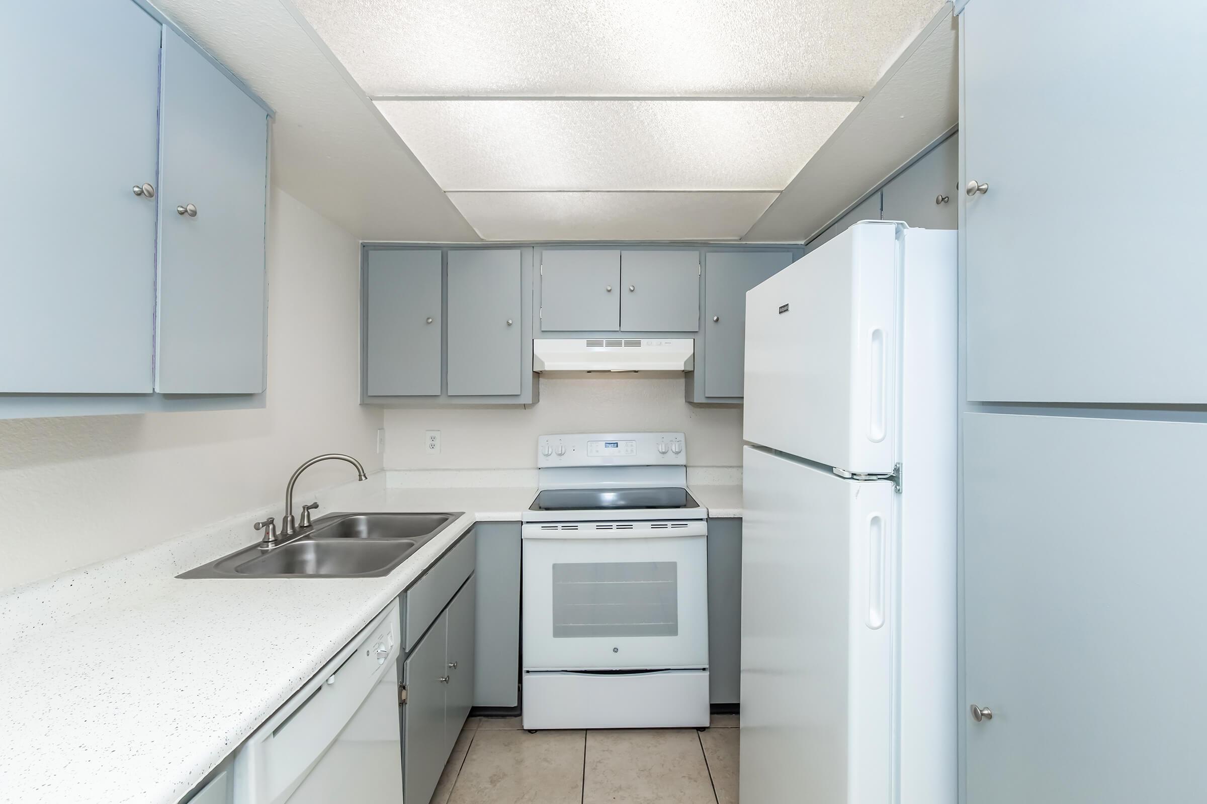 A well-lit kitchen featuring light gray cabinets, a white refrigerator, a white stove with an oven, a double sink, and neutral-colored tile flooring. The ceiling has fluorescent lighting, providing bright illumination throughout the space.