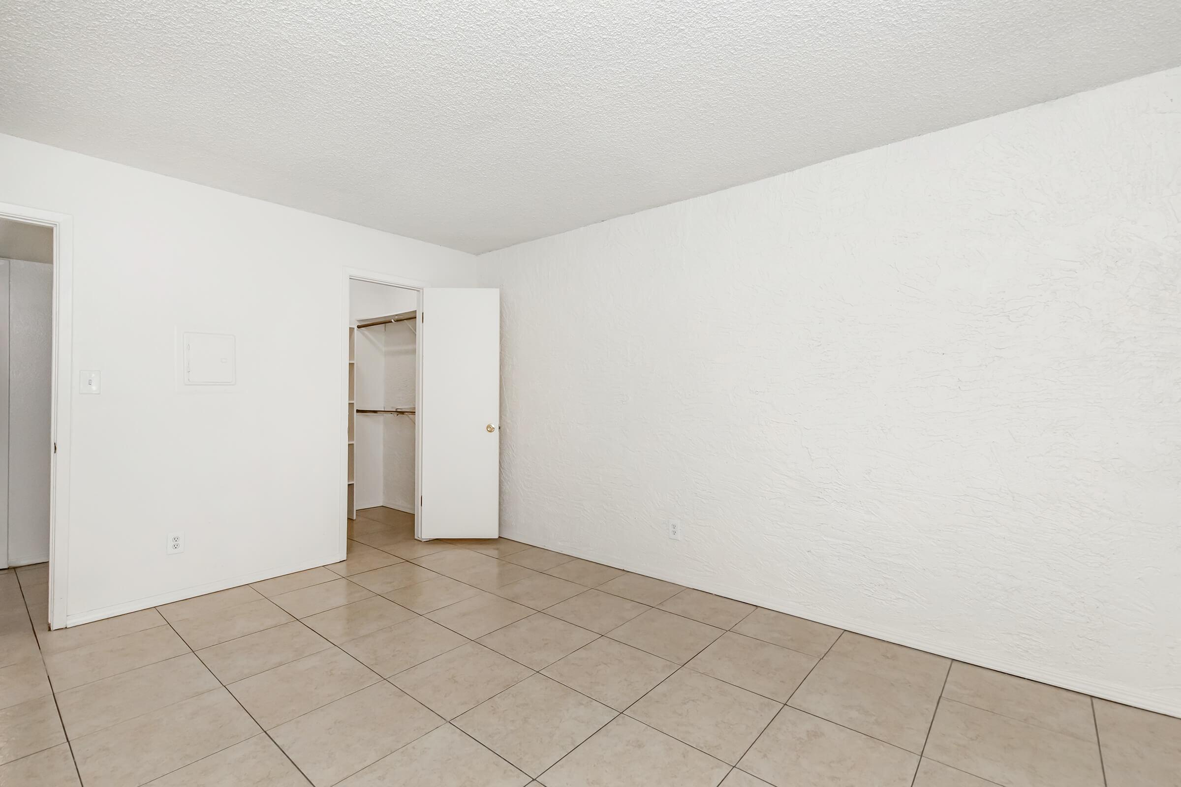 Empty room with white textured walls and tiled floor. A doorway on the left leads to a closet, and an open door on the right suggests access to another space. The room is well-lit, with no furniture present, creating a spacious and clean appearance.
