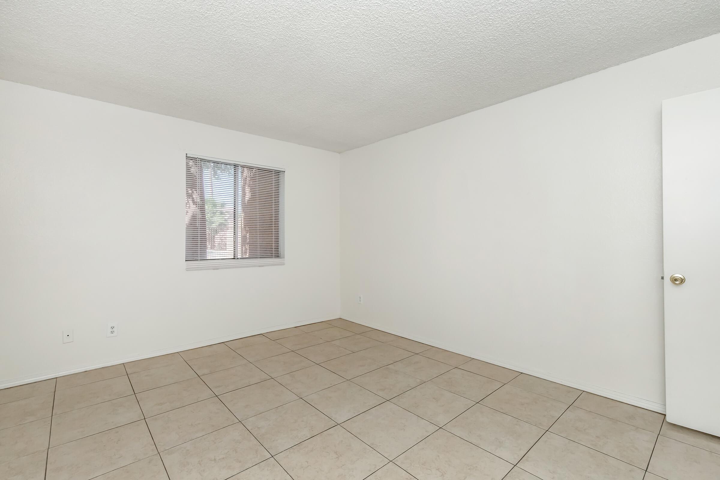 Empty room with white walls and tiled floor. A window with blinds is visible, allowing natural light to enter. The space appears clean and unfurnished, providing a neutral environment. A door is seen on the right side, leading to another area.
