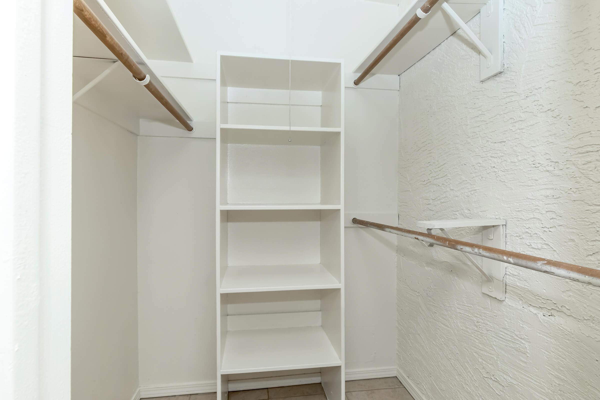 A spacious, empty closet featuring white walls, two wooden hanging rods, and a tall white shelving unit. The floor is tiled, and the overall space is well-lit, creating an organized and minimalist appearance.