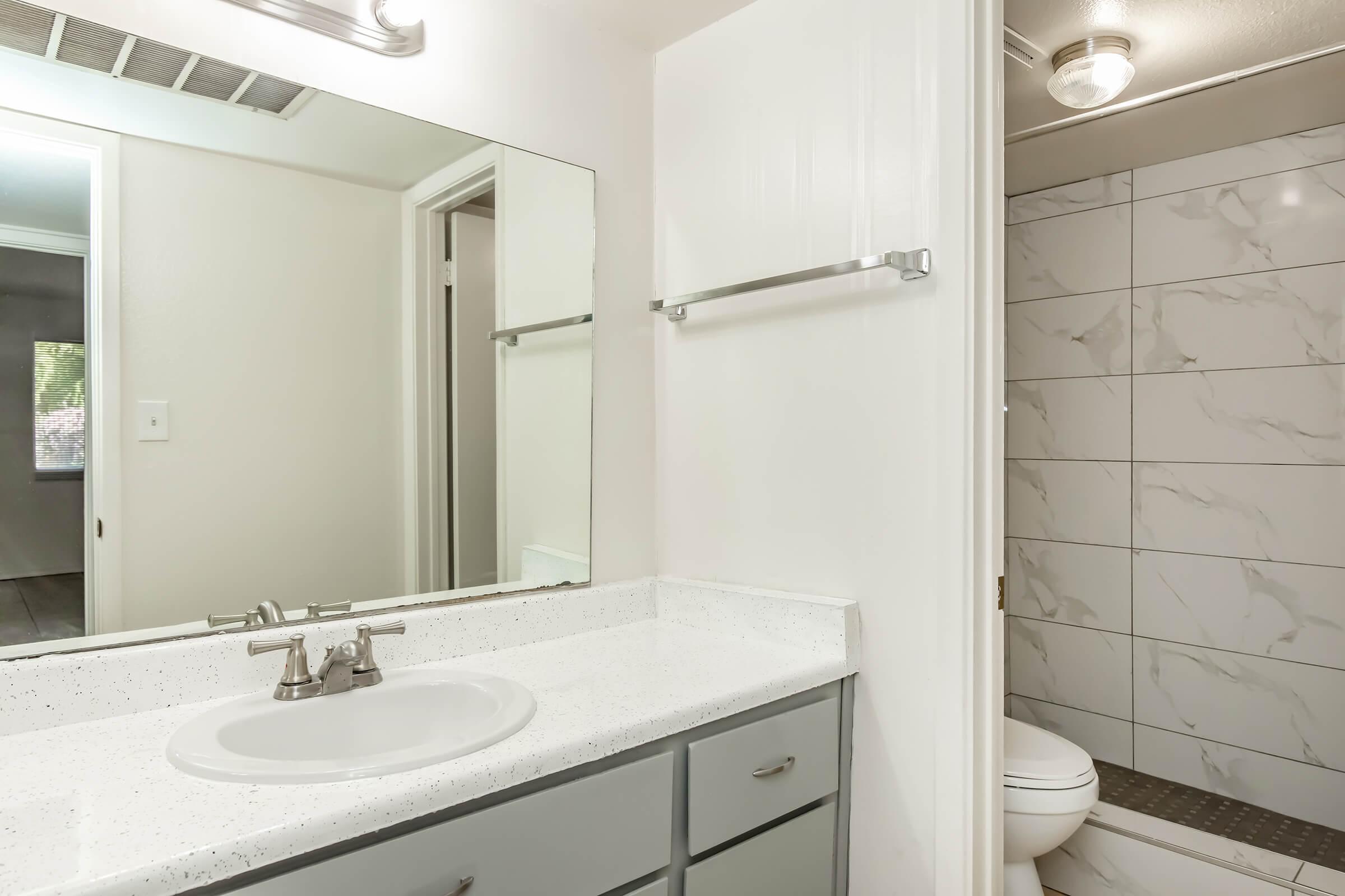 A bathroom featuring a white countertop with a sink, a large mirror above it, and gray cabinets. In the background, there is a shower area with white tiled walls and a toilet. The room is well-lit with natural light coming from a window.