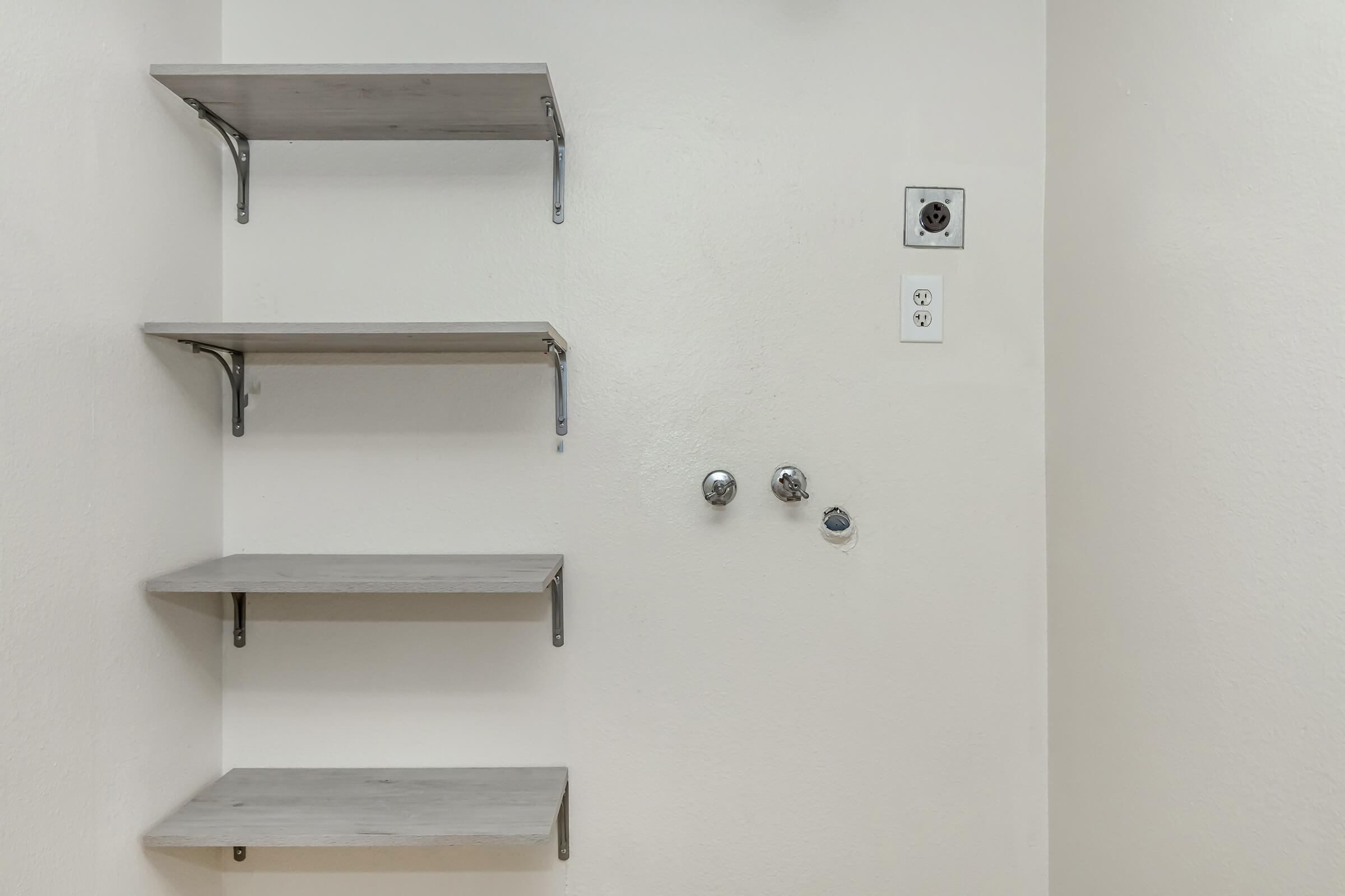 A plain wall with four wooden shelves mounted on brackets. There are two visible wall outlets and two plumbing fixtures on the right side. The shelves are empty, and the wall is painted a light color.