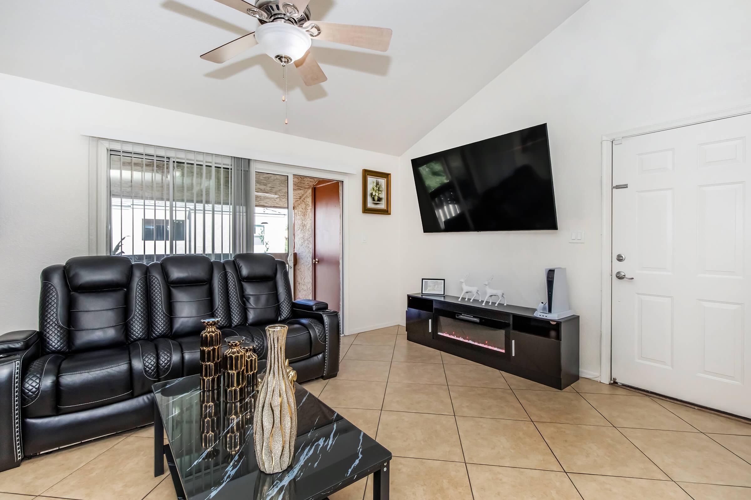Living room featuring black leather recliner chairs, a modern coffee table with decorative vases, and a wall-mounted flat-screen TV. A door leads to an exterior patio, and large sliding glass doors allow natural light to enter. Light-colored walls and tiled flooring enhance the space's openness.