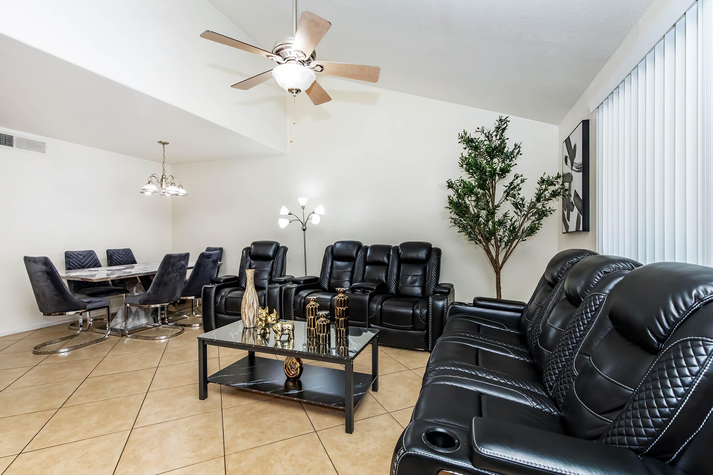 A modern living room featuring black leather recliners, a glass coffee table with decorative items, and a tall indoor plant. A dining area with a round table and elegant chairs is visible in the background. The space has light-colored walls and a ceiling fan, creating a stylish and inviting atmosphere.