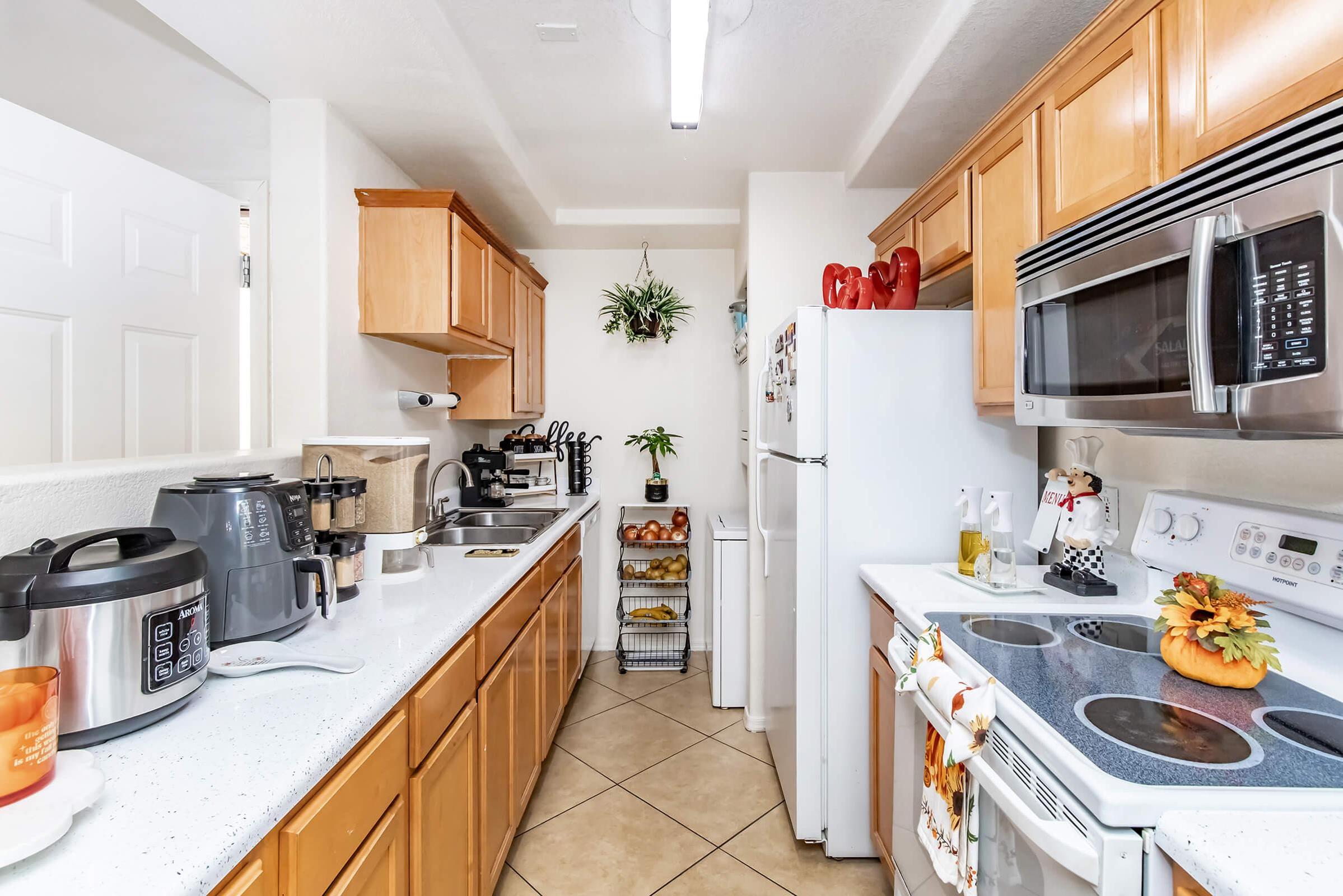 A modern kitchen featuring wooden cabinets, a white countertop, and stainless steel appliances. There is a refrigerator, stove, and microwave. Counter space includes a coffee maker and a slow cooker, with decorative plants and a pumpkin centerpiece adding a cozy touch.