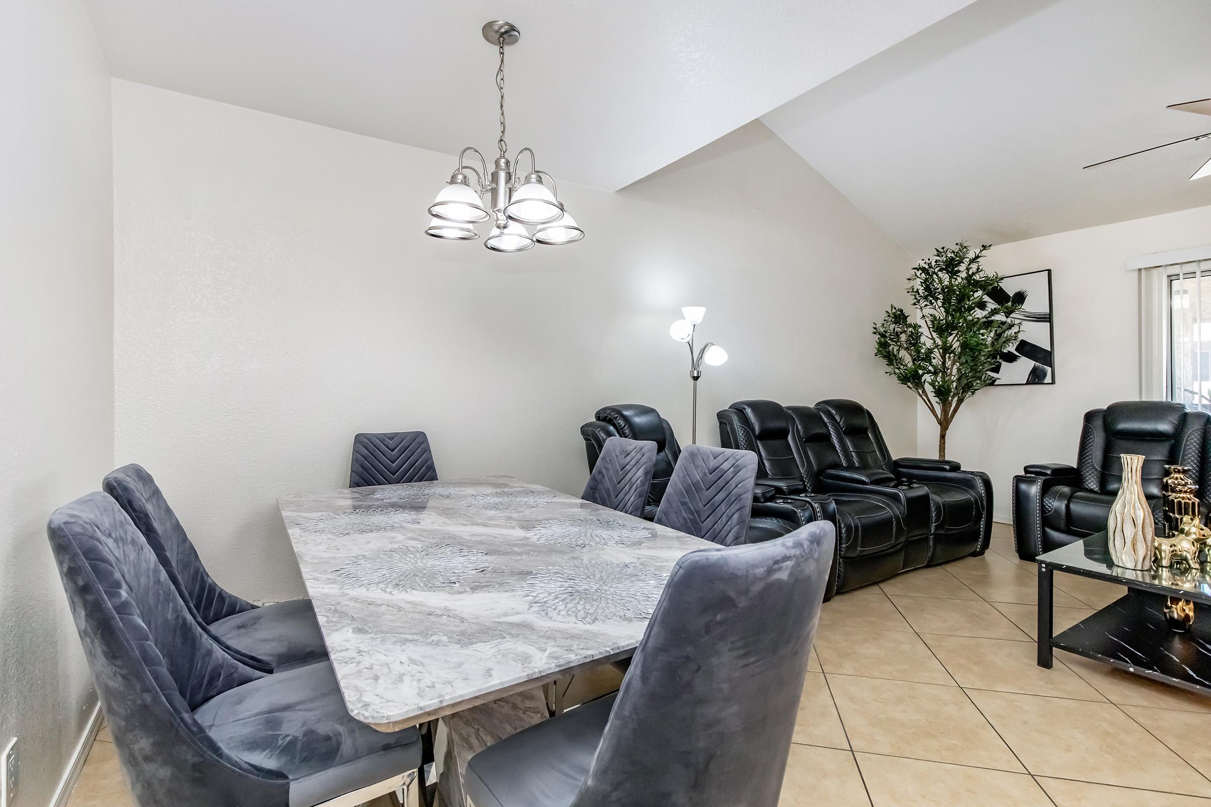 A modern dining area featuring a marble table surrounded by six velvet chairs. In the background, there are comfortable black leather recliners and a floor lamp. A decorative plant adds a touch of greenery, and the room has light-colored walls and tiled flooring.