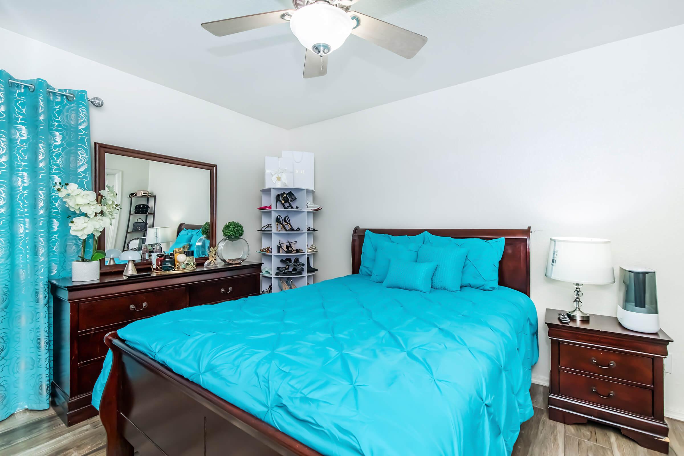 A bright bedroom featuring a large wooden bed with a turquoise comforter and matching pillows. There is a wooden dresser and a bedside table with a lamp. A mirror and shoe rack are also present, along with decorative items like flowers and a green plant. The walls are painted in a light color, enhancing the room's brightness.