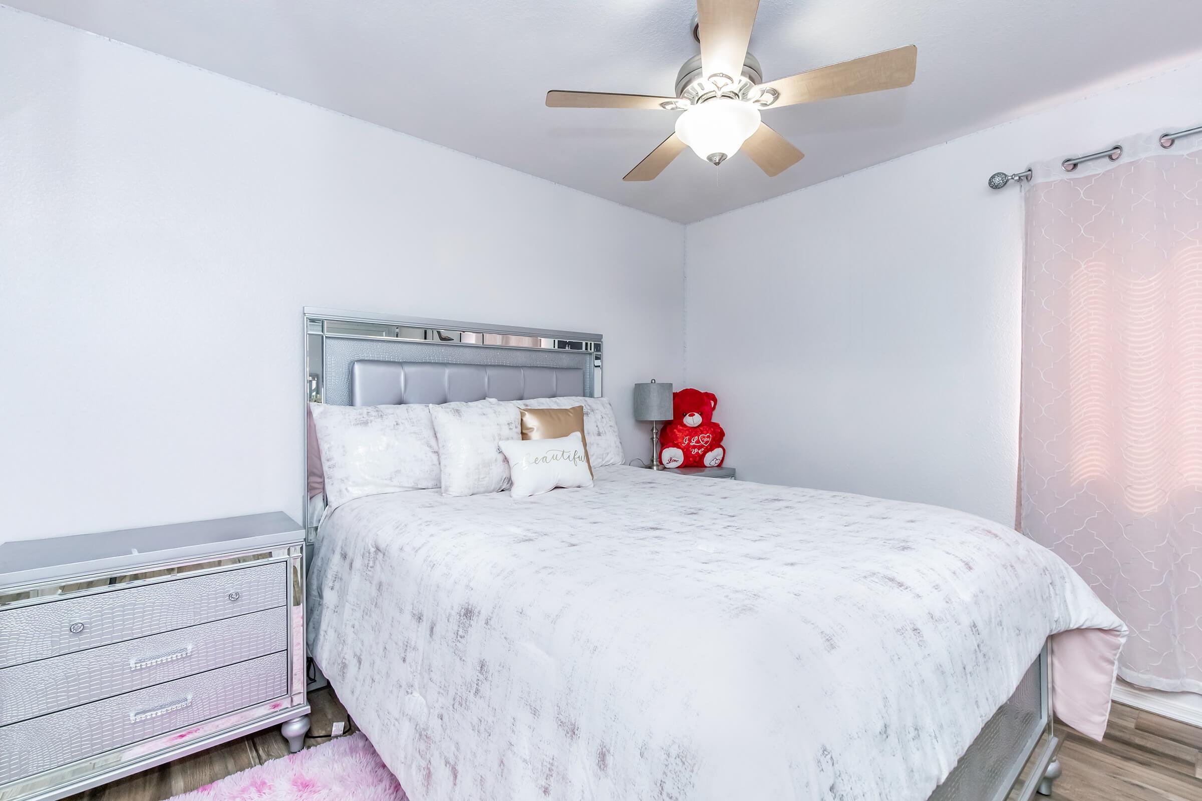 A cozy bedroom featuring a plush queen-sized bed with a light gray and white bedding set. A silver nightstand sits beside the bed, and a cute red teddy bear is perched on the bed. The room has soft pink curtains, a ceiling fan, and light-colored wood flooring, creating a warm and inviting atmosphere.