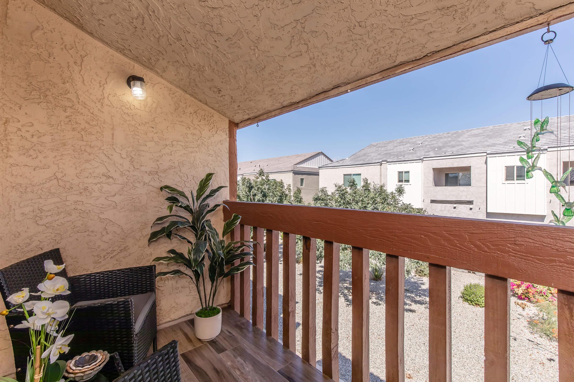 A cozy balcony with two wicker chairs and a small table, adorned with a vase of flowers. The view shows a landscaped area with shrubs and nearby buildings under a clear blue sky, creating a peaceful outdoor setting.