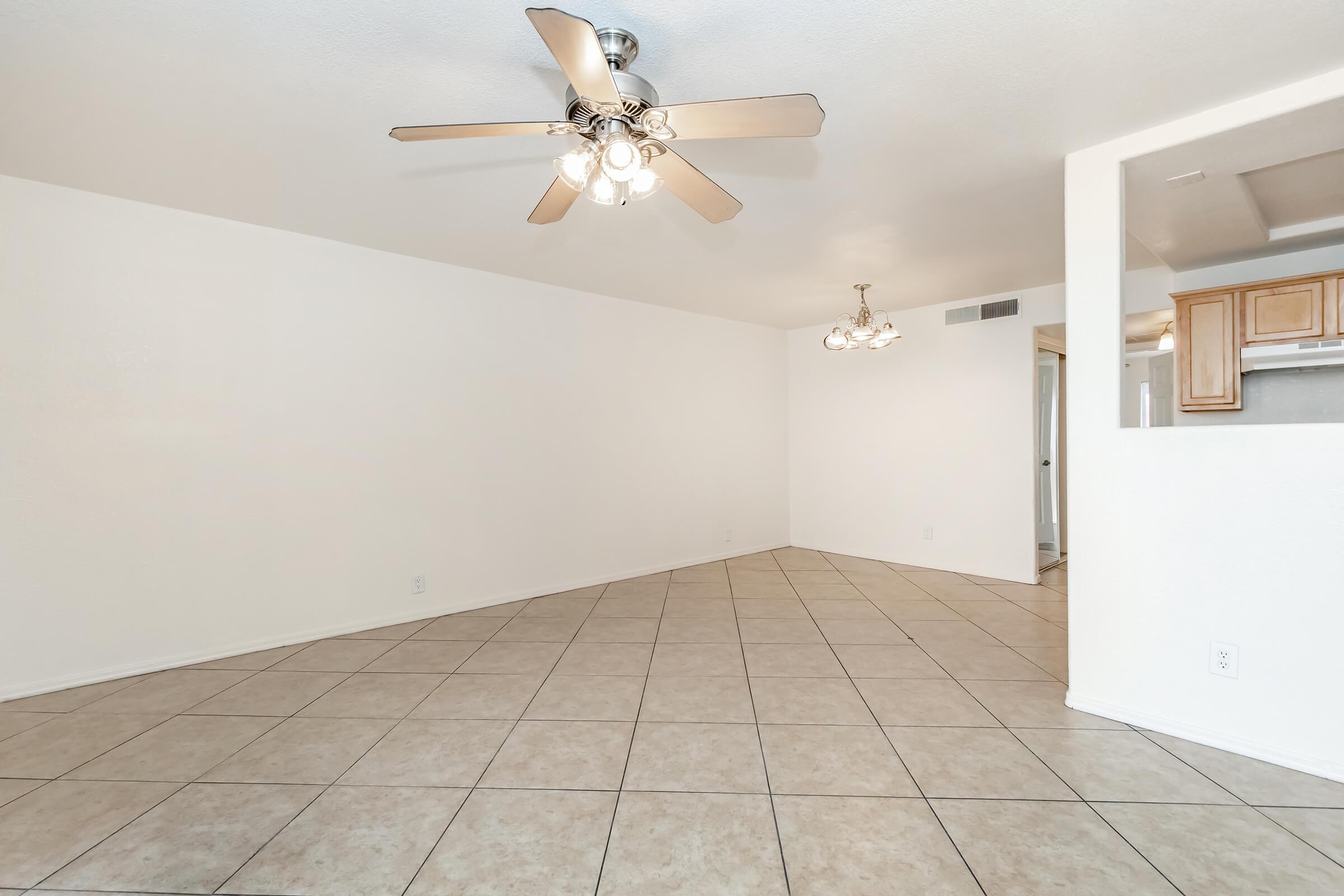 A spacious, empty living room featuring light-colored walls and beige tile flooring. A ceiling fan with light fixtures hangs in the center, and there are doorways leading to other rooms. Natural light fills the area, creating a bright and inviting atmosphere.