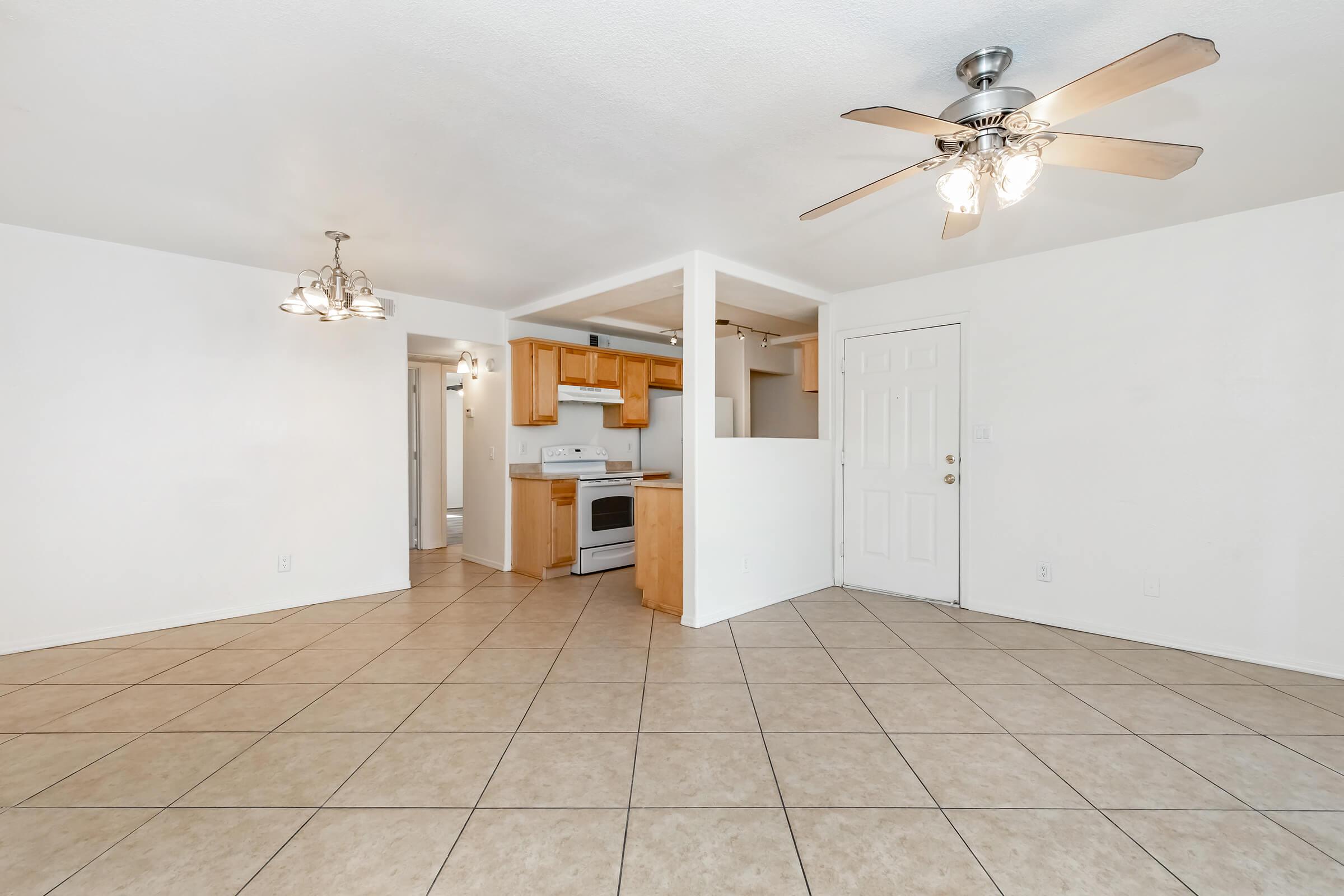 Spacious living area featuring beige tiled flooring, a ceiling fan with lights, and a simple layout. The kitchen is partially visible, with wooden cabinets and a white stove. The walls are painted white, enhancing the bright and airy atmosphere. Natural light fills the space from multiple sources.