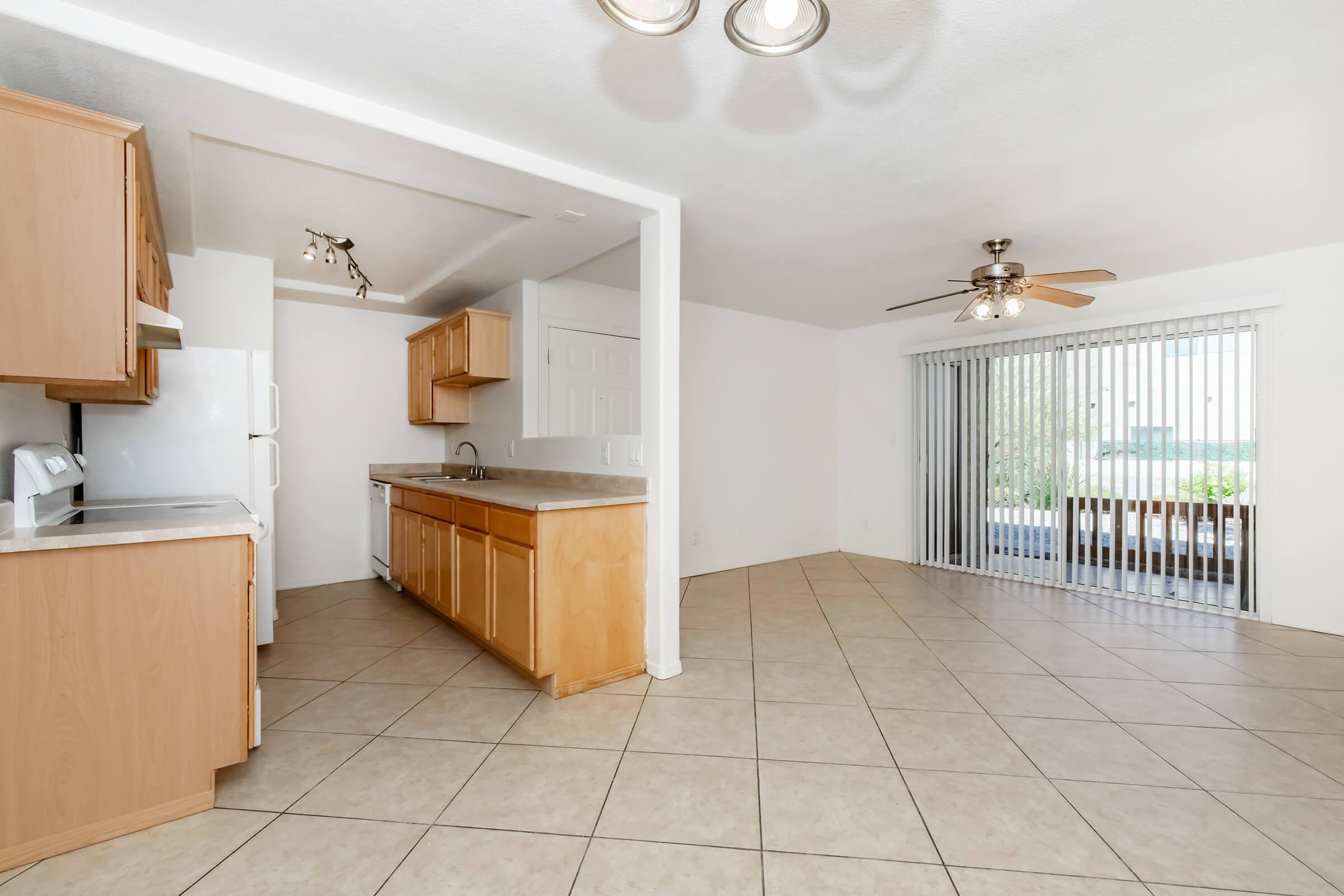 A spacious, well-lit kitchen and dining area featuring beige cabinets, a white refrigerator, and tile flooring. A sliding glass door leads to a patio area, with natural light illuminating the room. The ceiling has modern light fixtures and a ceiling fan.