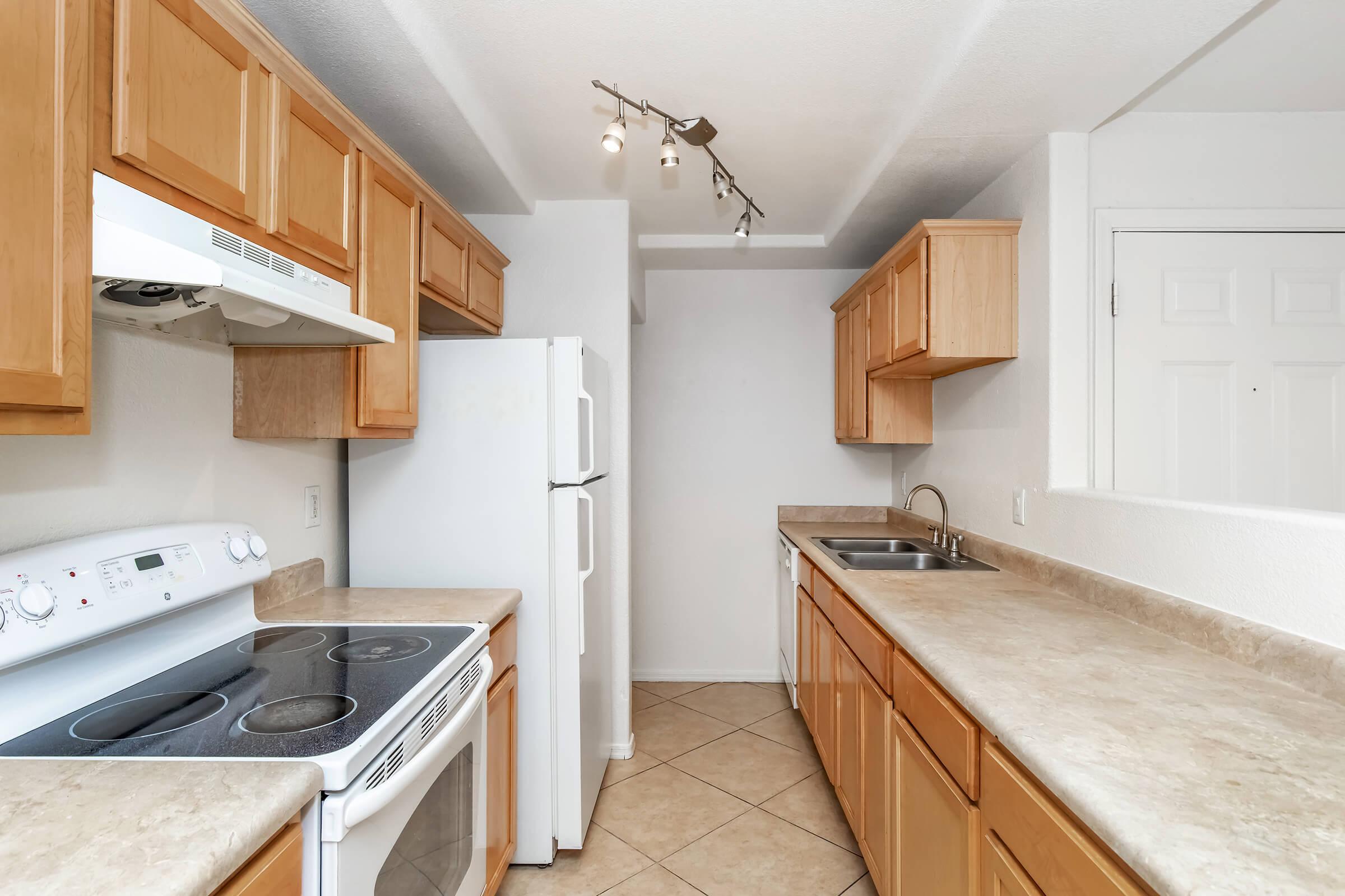A modern kitchen featuring light wood cabinetry, a white refrigerator, a white stove with an oven, and a stainless steel sink. The countertops are beige, and the floor is tiled. Track lighting illuminates the space, which has white walls and a door at the far end.