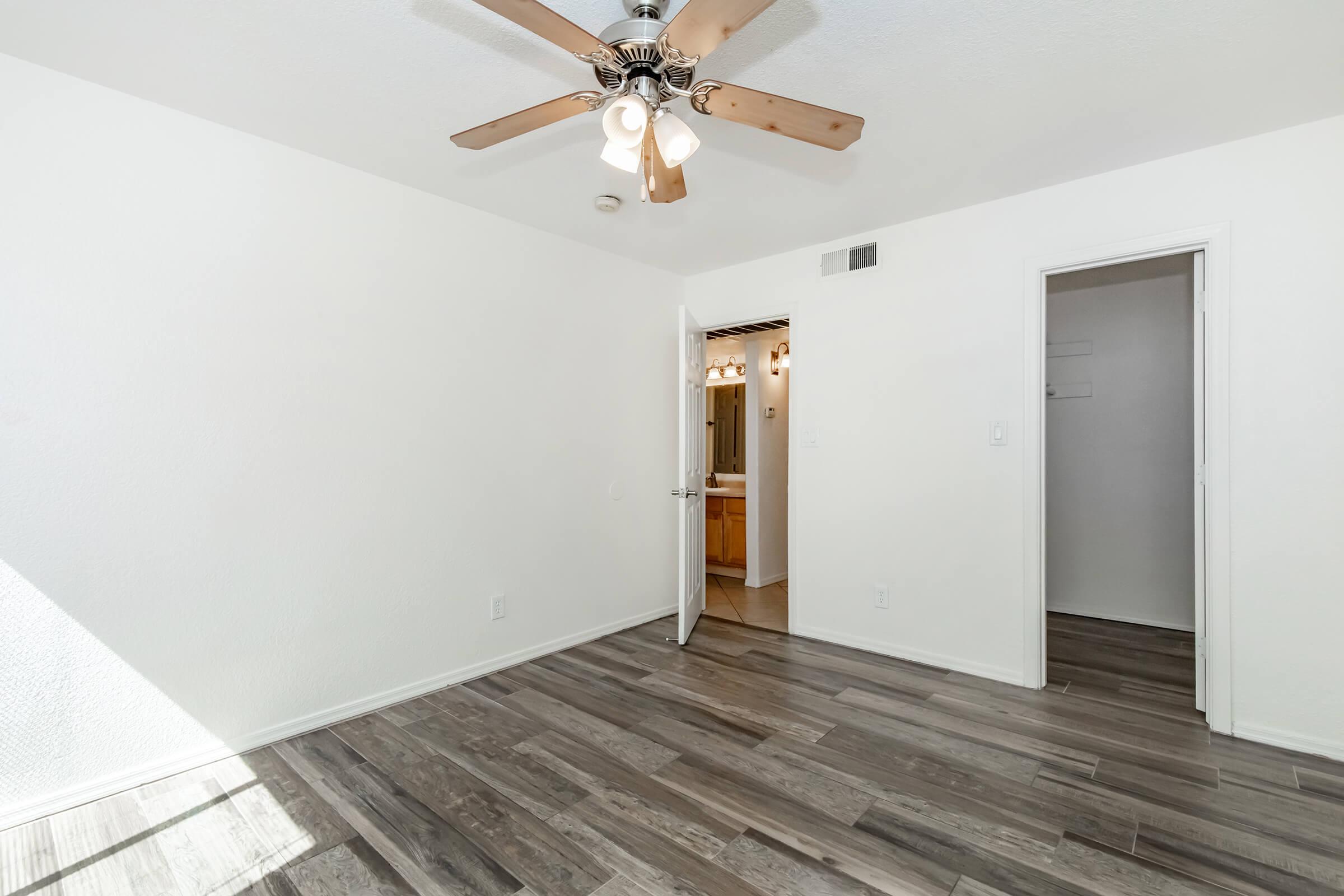 Interior view of a bright room featuring a ceiling fan with wooden blades, light-colored walls, and grey laminate flooring. A doorway leads to a bathroom, and there is a closet visible on the right. Natural light streams in, enhancing the spacious feel of the area.