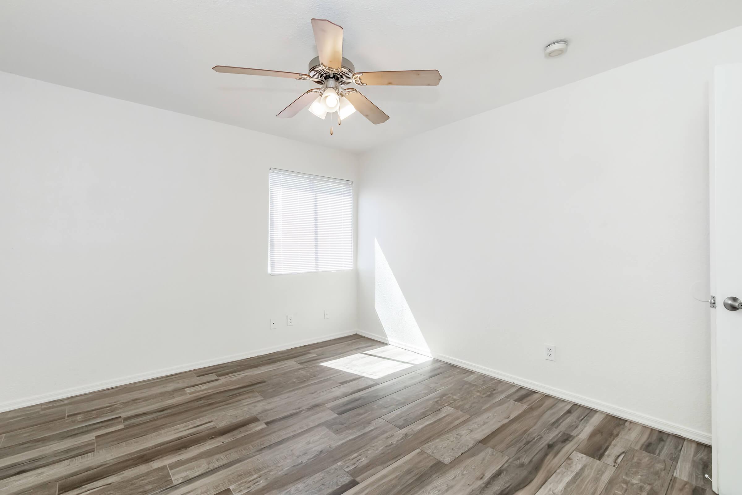 A bright, empty room with a ceiling fan, light-colored walls, and wooden flooring. A small window allows natural light to enter, creating a spacious and inviting atmosphere.