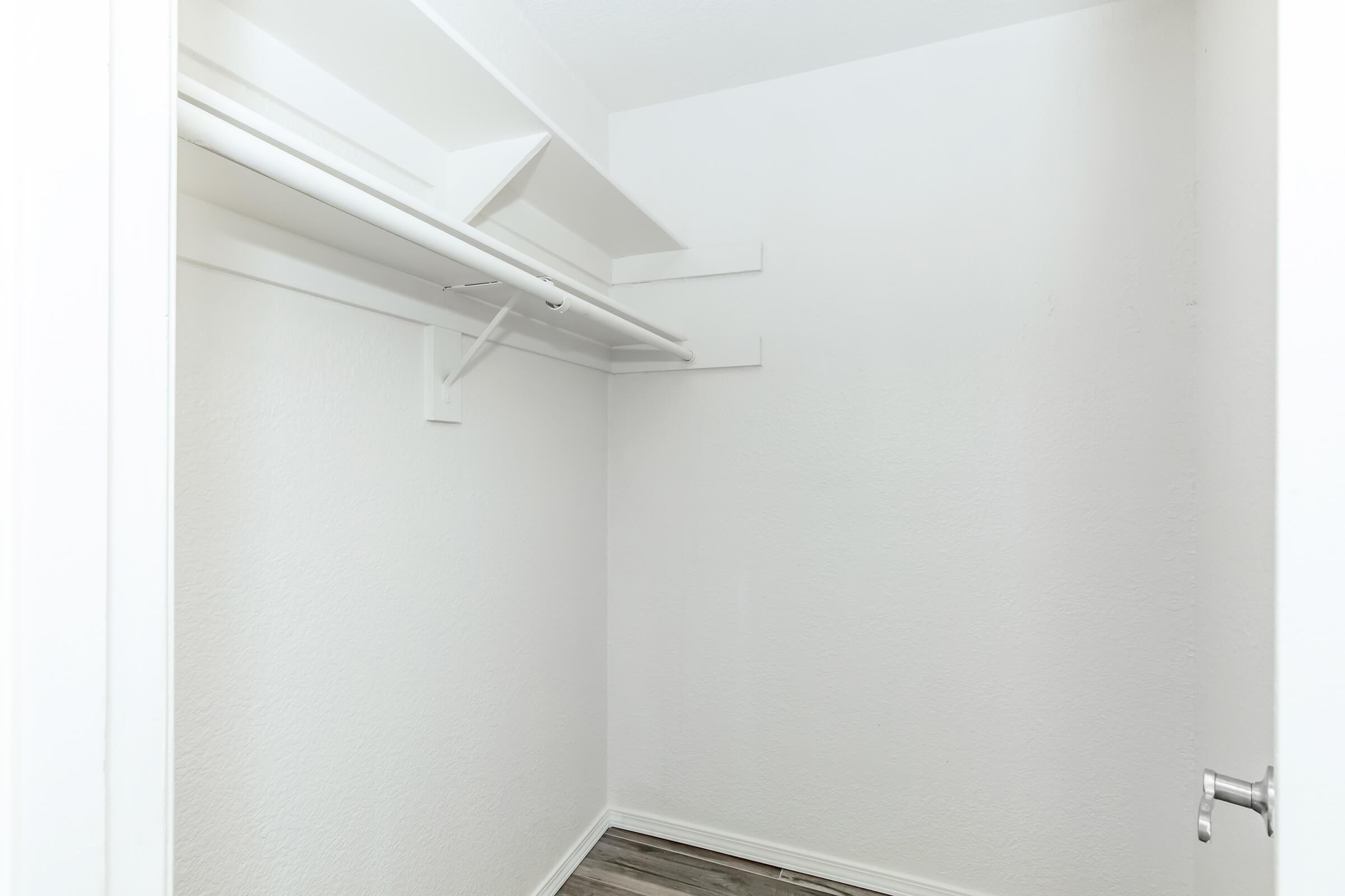 A small, empty closet with white walls and a wooden floor. The closet features a clothing rod for hanging items, and there are no shelves or storage bins visible. The overall space is clean and uncluttered.