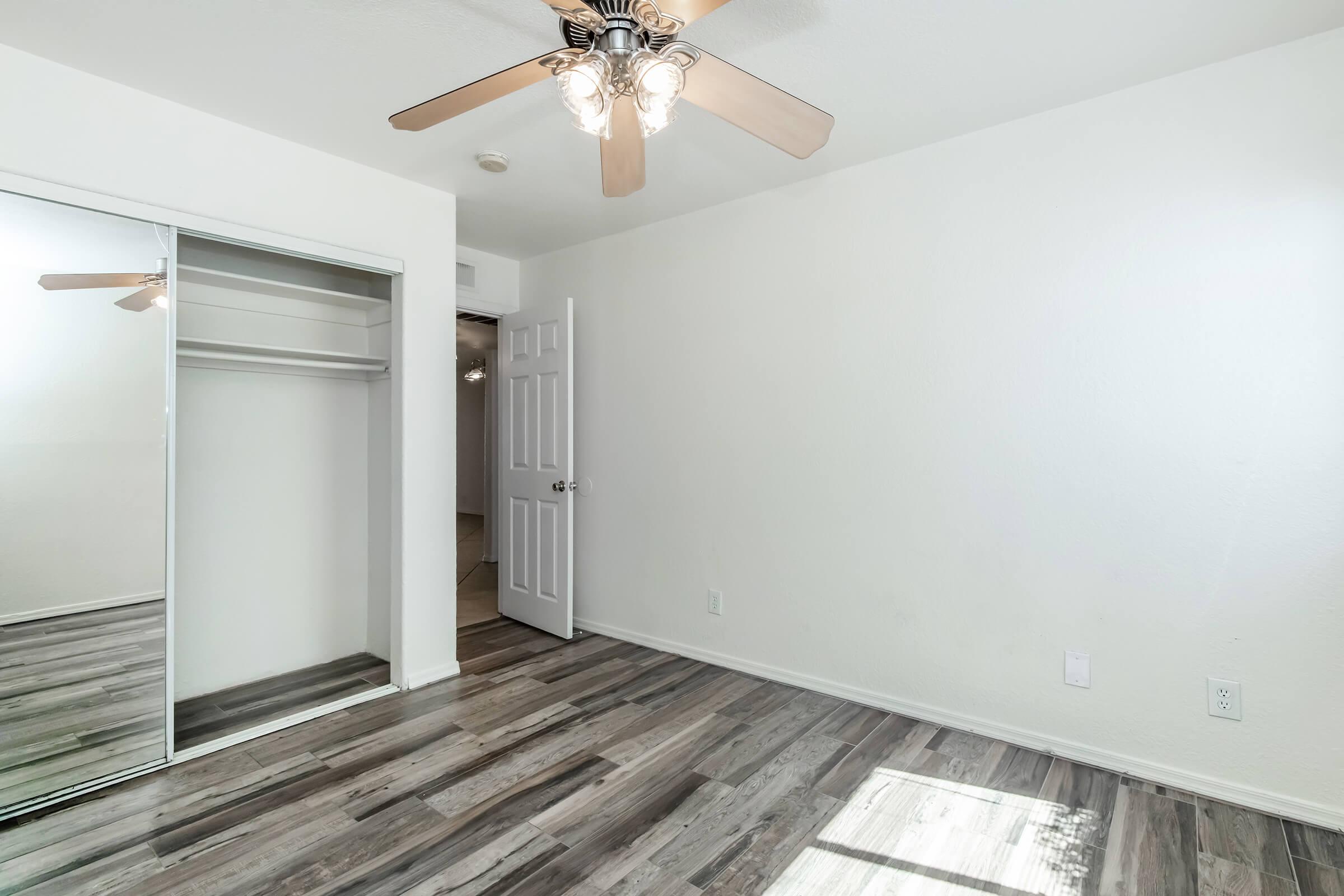An empty room with light-colored walls and wooden flooring. It features a ceiling fan, a mirrored closet door, and an open doorway leading to another space. The room is well-lit by natural light coming in through the doorway.