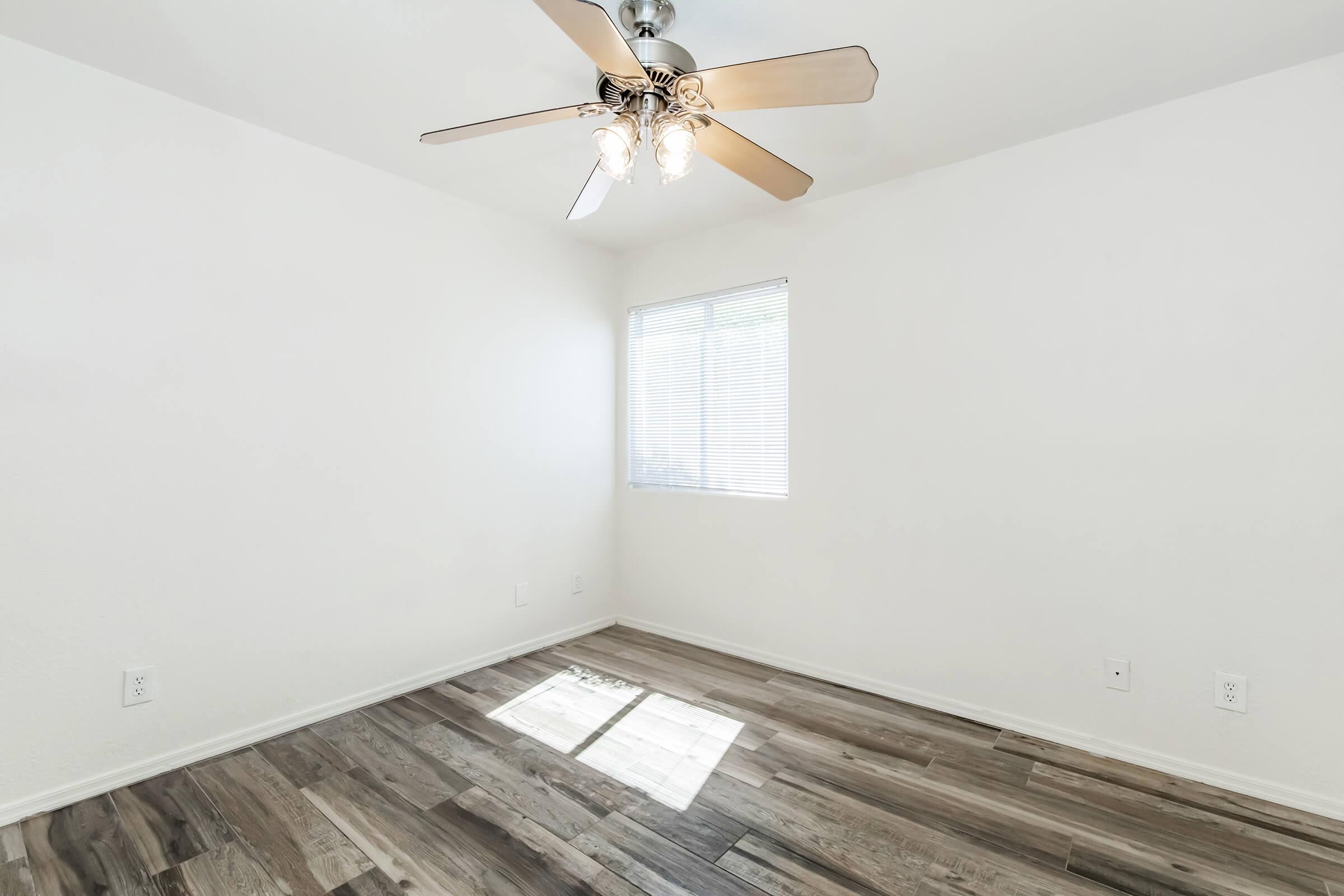 A well-lit empty room with light-colored walls and a ceiling fan. The floor features wood-like laminate flooring. A window with blinds allows natural light to enter, creating a bright atmosphere. The space is unadorned and ready for furniture placement.
