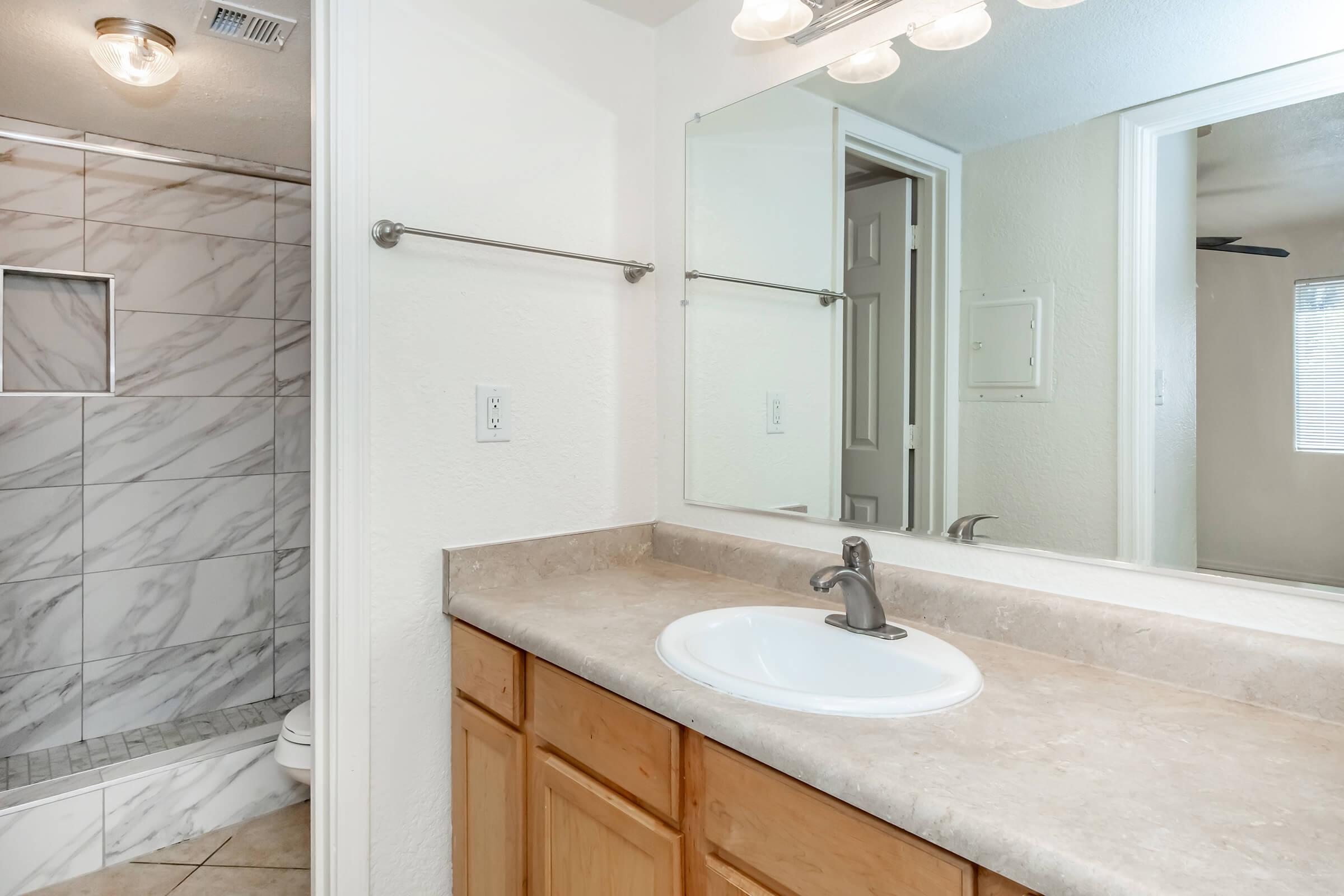 A modern bathroom featuring a marble-tiled walk-in shower with a glass door. There is a single sink with a chrome faucet on a light-colored countertop and wooden cabinetry. A mirror is mounted above the sink, and a doorway leads to another room, with natural light coming in from a window.